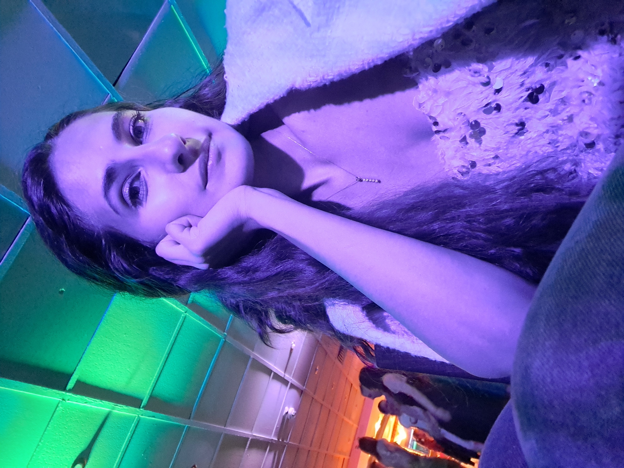 The image is a close-up, selfie-style portrait of an adult female, illuminated primarily by artificial purple light. The woman, positioned in the lower left quadrant, has long, dark, wavy hair and is looking directly at the camera. Her left hand is resting with fingers under her chin, forming a relaxed pose. Her face shows distinct eye makeup, including eyeliner and possibly glitter on the eyelids. She wears a light-colored, possibly white or cream-colored, textured jacket or shawl draped over her right shoulder. Beneath this, a light-colored, possibly sequined or embellished top is visible on her chest. A delicate gold-colored necklace with a small, vertical pendant adorns her neck. The lower part of the frame shows a segment of dark-colored denim fabric, likely part of her attire.
The background features a ceiling with a grid-like panel structure, illuminated by varying colored lights. The upper left portion of the ceiling displays green and blue illumination, transitioning to warmer, orange-pink tones on the right side. Several indistinct figures of other people are visible in the blurred background on the right, suggesting a public or social gathering place. No discernible features within the image provide information regarding its specific geographical location, including the city of Sofia, Bulgaria.
