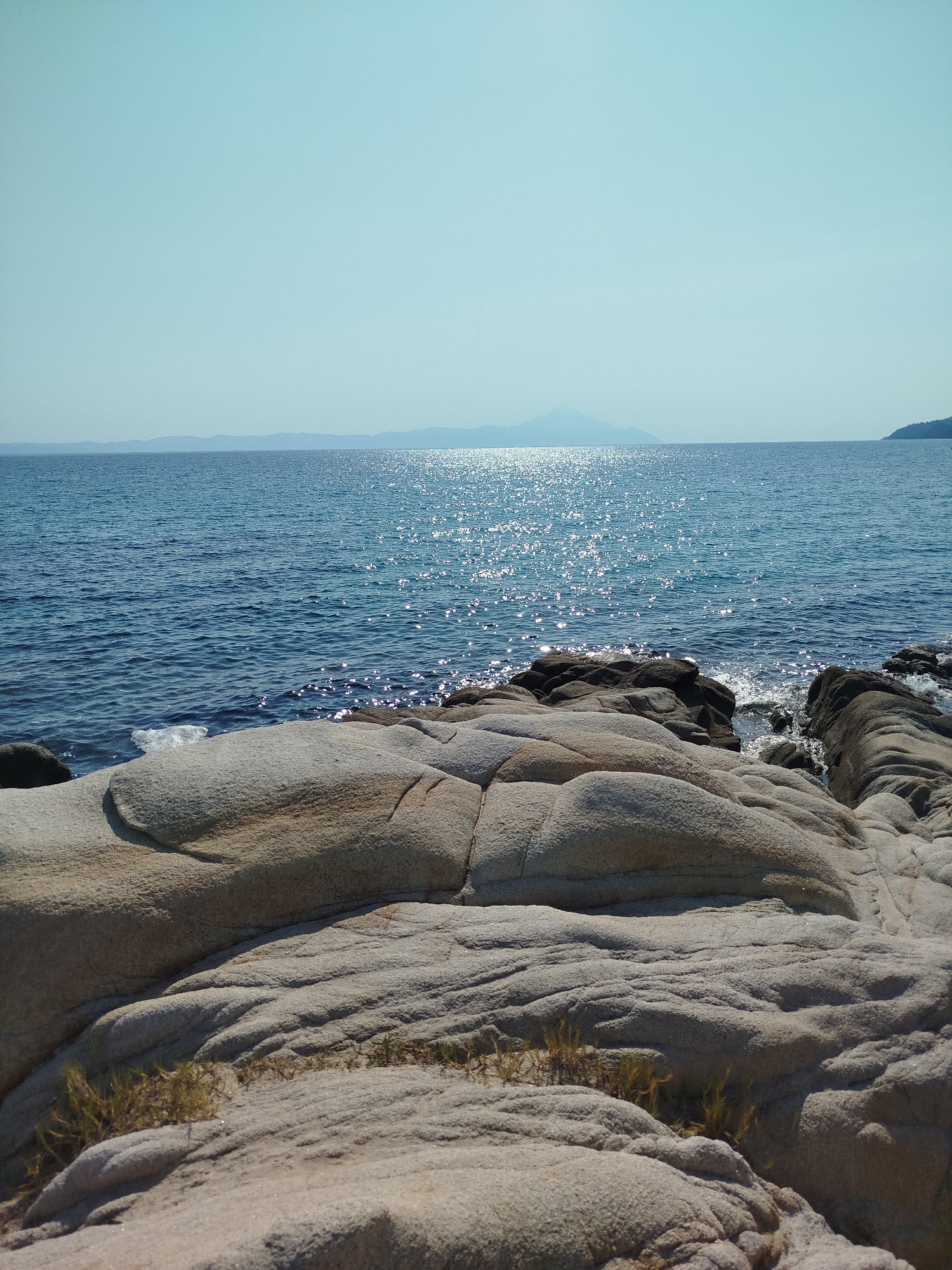 The image depicts a serene coastal landscape on a sunny day. The foreground is dominated by large, smooth, light-colored rocks, worn by the elements, with patches of dry grass growing between them. These rocks form a natural, textured barrier leading towards the sea. The water is a deep blue, reflecting the bright sunlight with a shimmering, sparkling effect. In the distance, a hazy mountain range is visible on the horizon, silhouetted against a pale, clear sky. The scene is one of natural beauty and tranquility, likely in Sithonia, Greece, known for its picturesque coastlines. There are no people or any man-made structures visible in the image.
