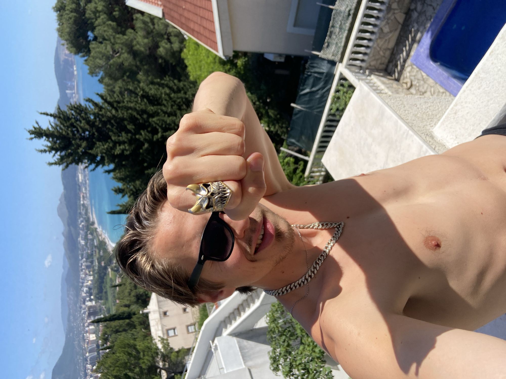 A shirtless male with light brown hair and sunglasses is taking a selfie in an outdoor setting. He is wearing a large ring and a silver chain necklace. The background features a cityscape with buildings, trees, a body of water, and a blue swimming pool. The location is most likely Šušanj, Montenegro, based on the typical Adriatic coastal geography and architecture.