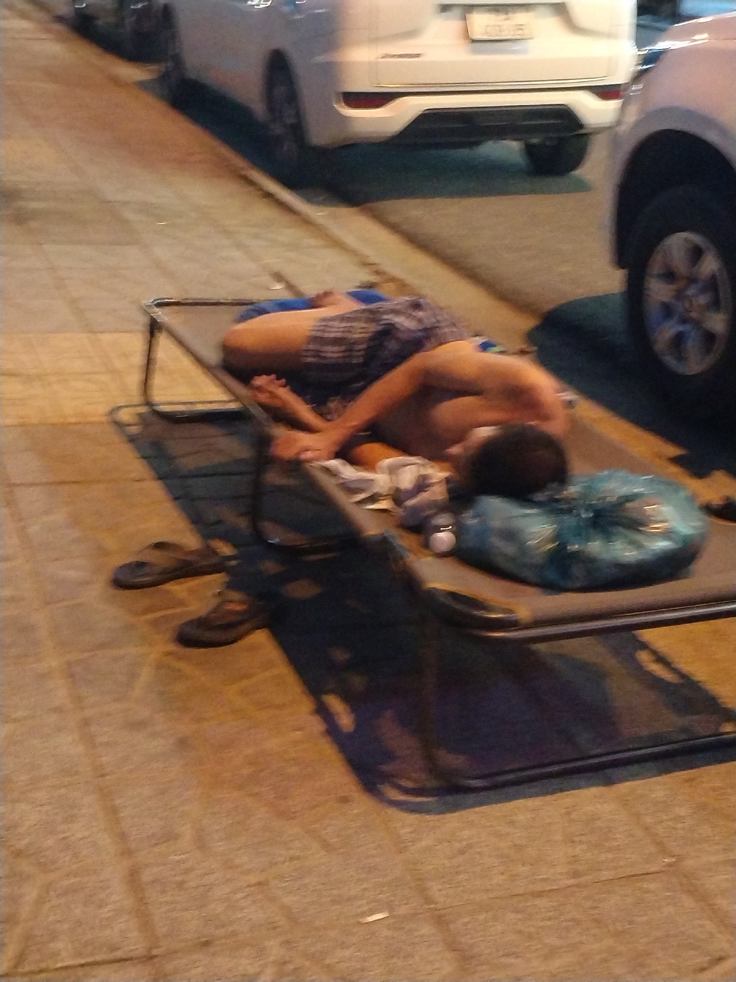A shirtless person wearing checkered shorts lies prone on a portable cot, situated on a tiled sidewalk adjacent to parked vehicles. Two pairs of footwear, including sandals and shoes, are placed near the cot. Bac Nha Trang Ward, Vietnam.
