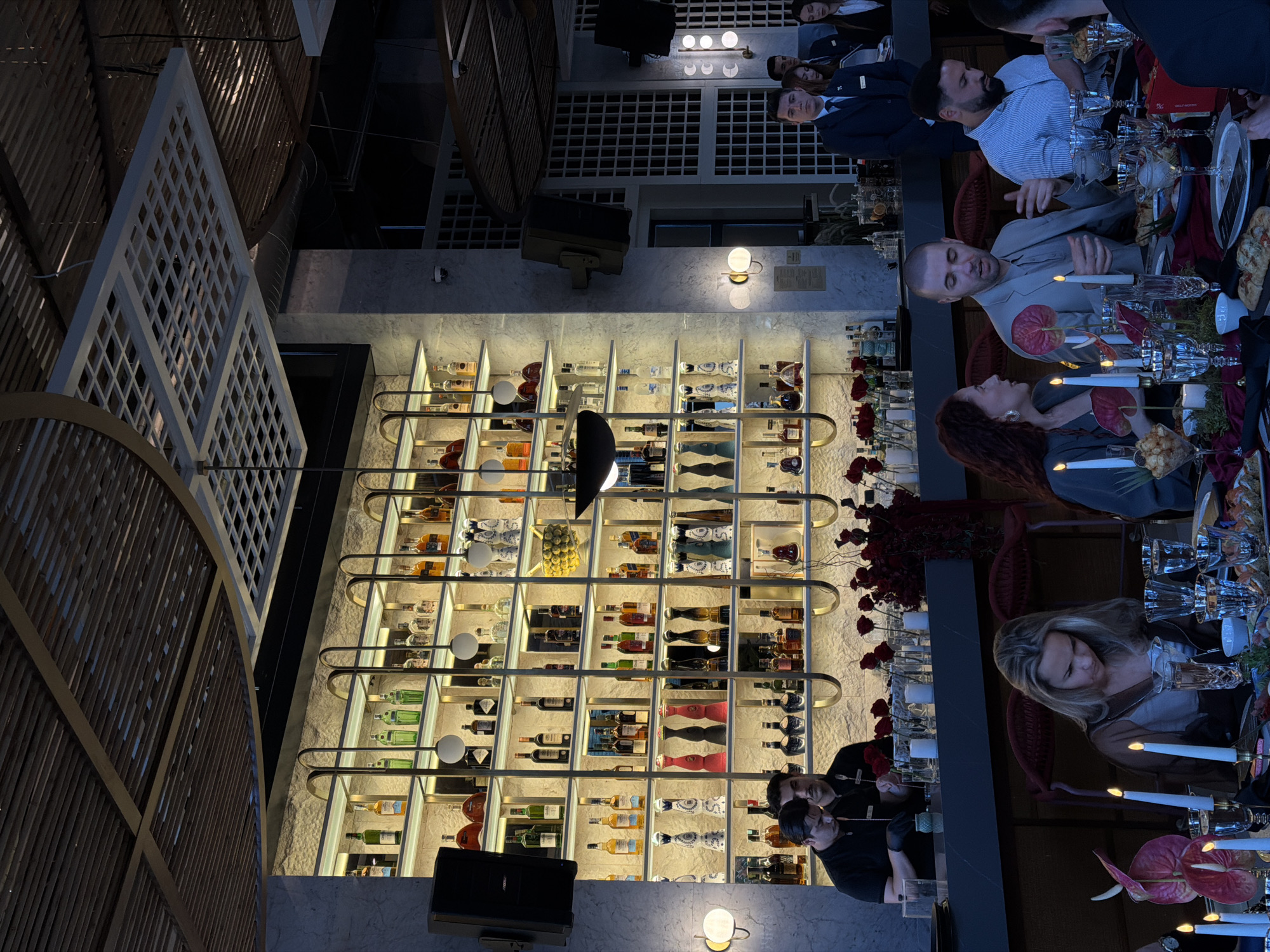 A tilted interior shot shows a restaurant scene in Budva, Montenegro. A wall-sized, illuminated display case holds a variety of bottled beverages. A dining table spans the right side of the image, occupied by several people engaged in conversation. The table is set with glassware, candles, and floral arrangements. The background features architectural elements including a slatted ceiling.