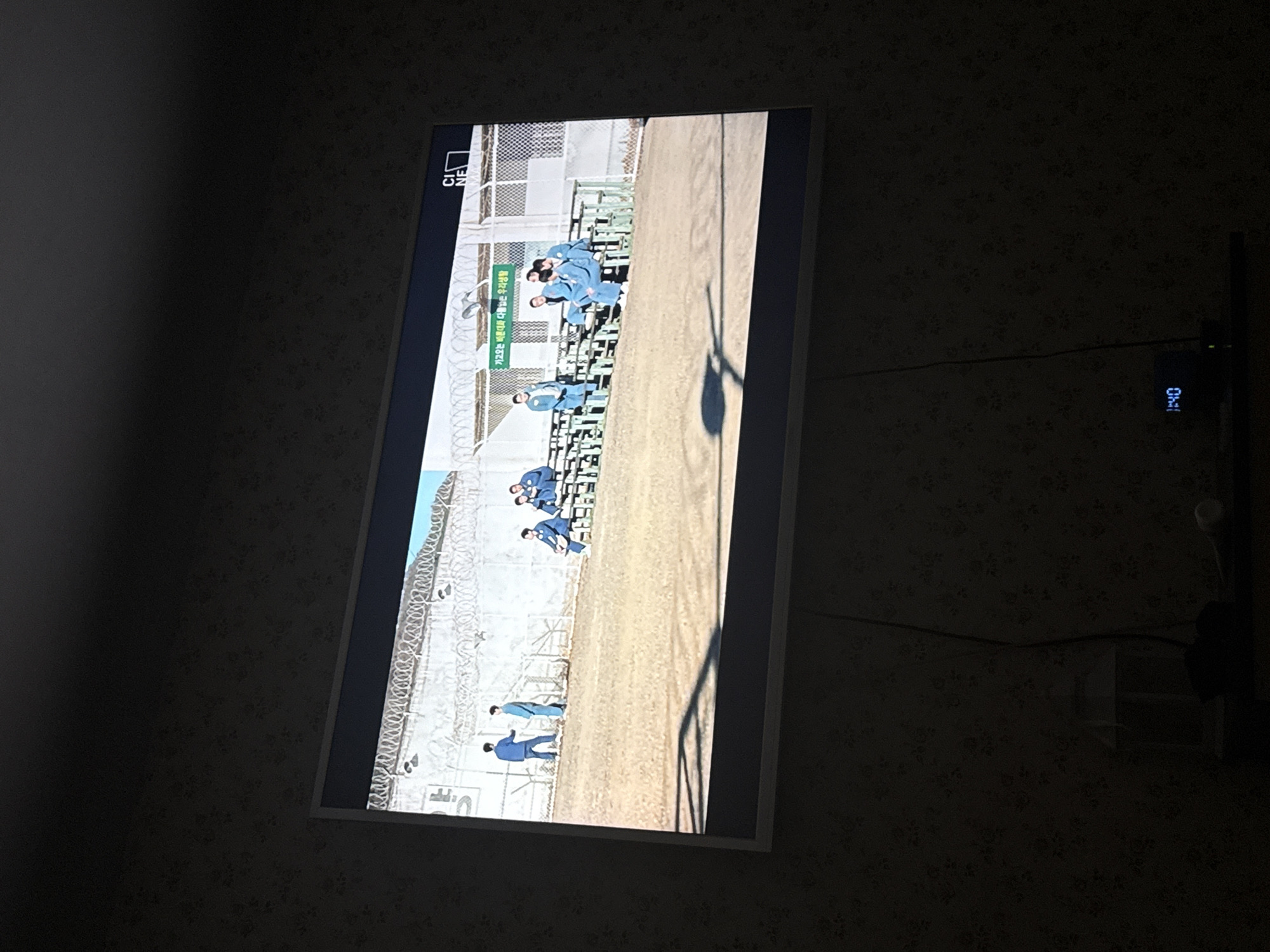 A television mounted on a wall displays a scene of uniformed figures, presumably prisoners, within a fenced and barbed-wire-topped enclosure. The location is not discernible within the visible content of the image. A digital clock next to the television indicates the time as "1:40." The room is dimly lit, with patterned wallpaper.