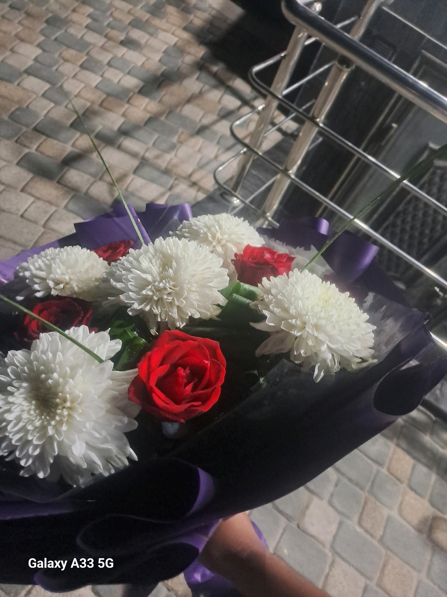A bouquet of white chrysanthemums and red roses is displayed in front of a railing and a paved ground. The bouquet is wrapped in dark purple paper. The scene is outdoors in Qorowul, Uzbekistan.