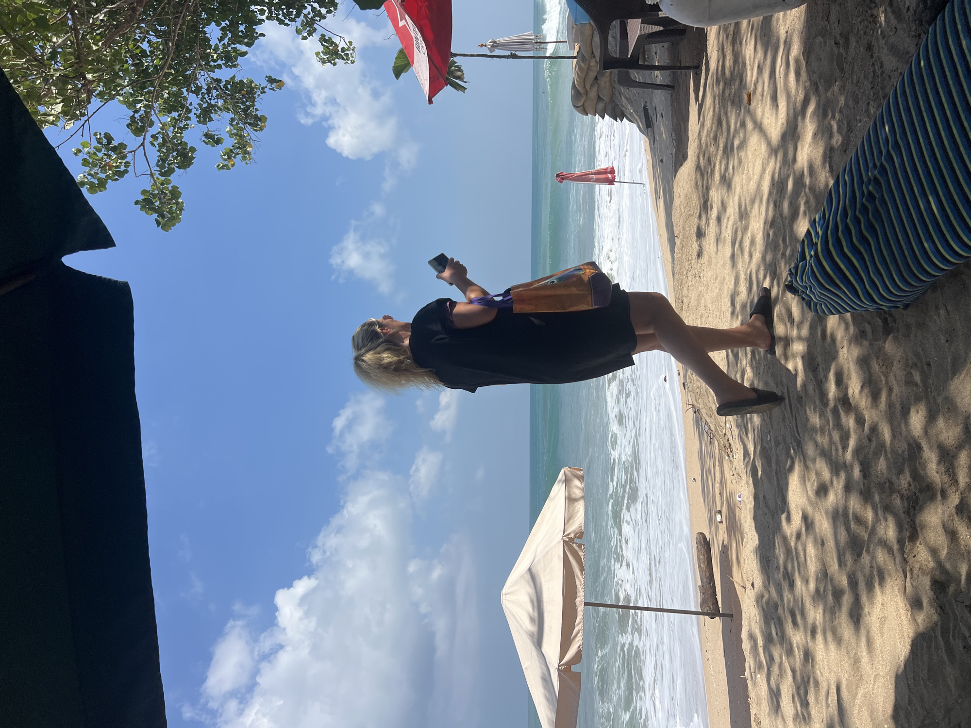 A woman with blonde hair, wearing a black dress and flip-flops, stands on a sandy beach in Kuta, Indonesia, holding a phone and a tote bag. She is facing the ocean, with waves rolling in. In the foreground, shadows from trees are cast on the sand. Various beach umbrellas and sandbags are visible, along with a piece of driftwood and some scattered trash. The sky is blue with scattered clouds.