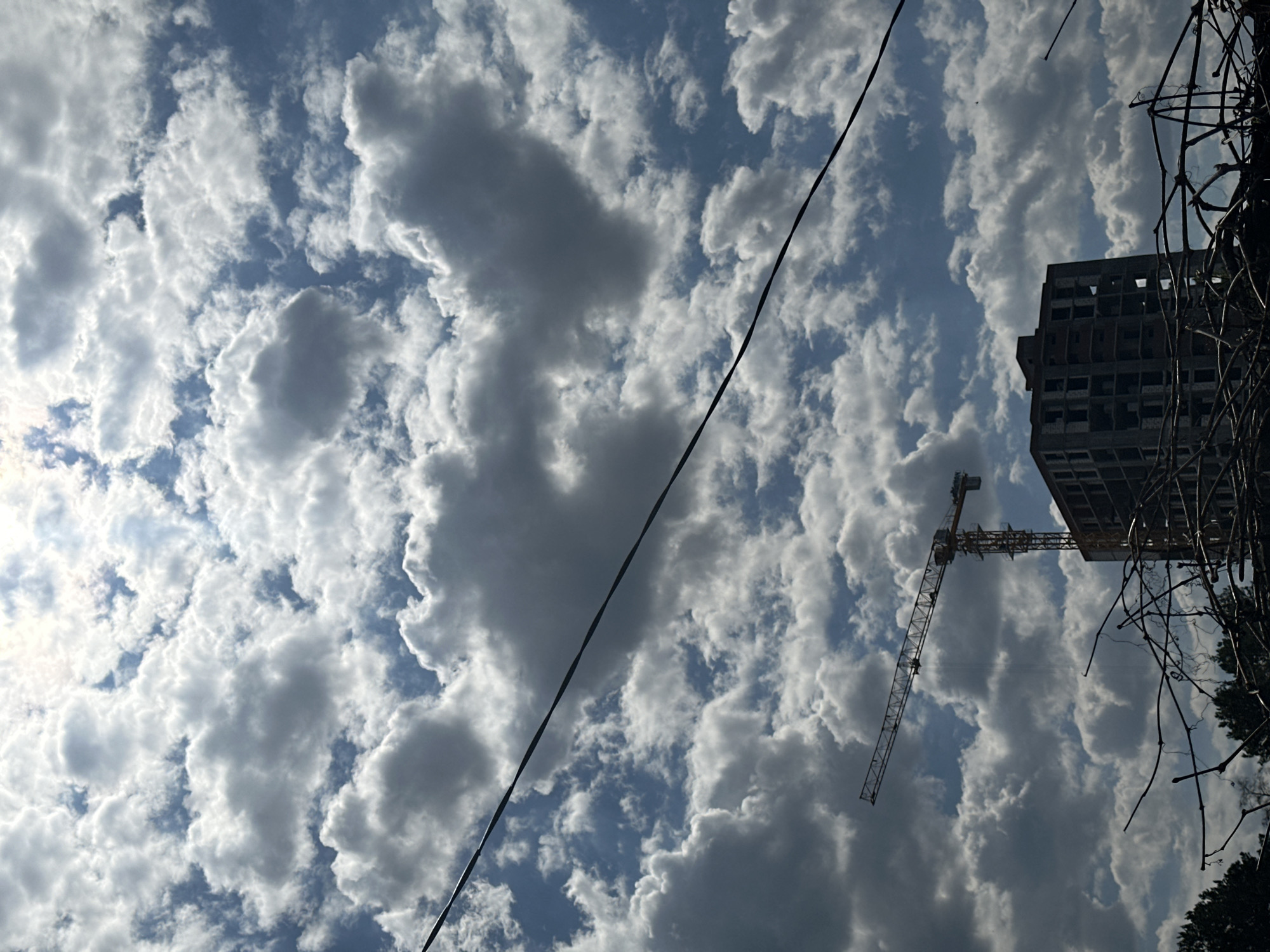 The image depicts a construction site in Chaikovsky, Russia, under a sky filled with cumulus clouds. A partially constructed high-rise building stands adjacent to a yellow construction crane. A power line stretches diagonally across the sky. Bare tree branches obscure parts of the building and crane.