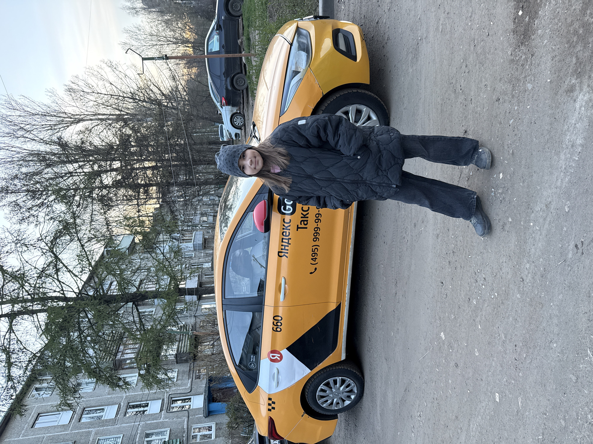 The image shows a young girl standing in front of a yellow Yandex Taxi car in Rusanovo, Russia. The girl is wearing a dark jacket, dark pants, and a gray hat. The taxi has the Yandex logo and phone number on its side. Several other cars are parked in the background. The scene appears to be on a street with a multi-story building and trees lining the sidewalk. The sky is overcast. The ground is paved.