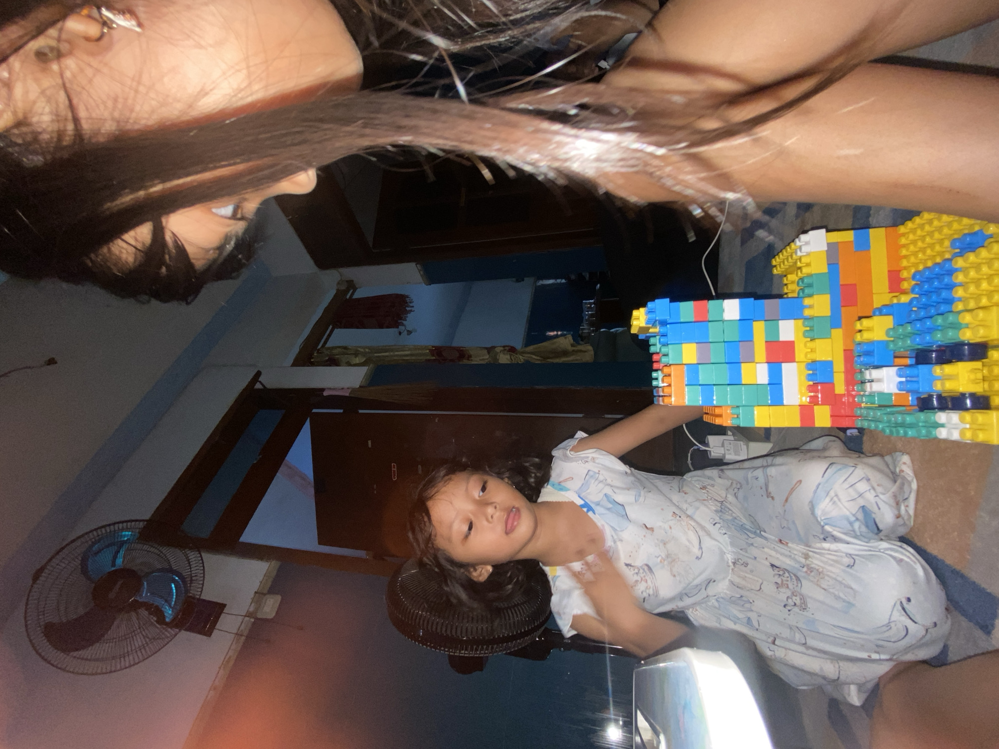 This image, taken in Gelam, Indonesia, captures an indoor scene, likely during the evening or night due to the dim, artificial lighting. In the central and lower part of the frame, a young child, appearing to be a girl, is seated on a patterned rug. She has dark hair and is wearing a light-colored, patterned dress or pajamas. She looks towards the viewer with a neutral expression, her mouth slightly agape, and one hand is near a large, colorful structure built from interlocking plastic blocks. This structure, a prominent feature in the image, is composed of various colored blocks including blue, yellow, red, green, white, and orange, forming what looks like a wall or tower.
Above the child, in the upper part of the frame, a person, presumably an adult female, is partially visible. Only her head, with long dark hair, and part of her bare arm are seen. She is looking down, seemingly at the child or the block creation. An earring is visible on her ear.
The background shows details of a home interior. A black electric fan is positioned on the floor near the child. Further back, a wall-mounted electric fan with blue blades is visible. Dark wooden furniture, possibly a cabinet or door, is also present. A power adapter is seen on the floor near the block structure. The overall impression is one of domestic activity, possibly playtime, in a modest setting. No discernible text is visible in the image.