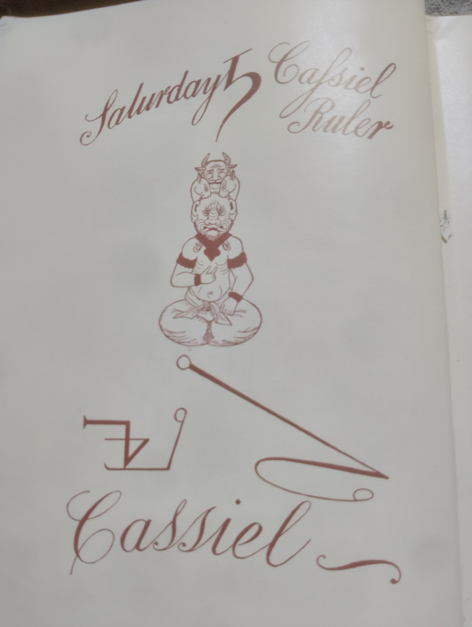 The image displays an antique-style illustration on a page. At the top, elegant script reads "Saturday 5 Cassiel Ruler". Below this text is a drawing of a stylized, multi-headed figure seated in a meditative pose, with horns and fierce facial features. Beneath the figure, a geometric symbol is drawn in a reddish-brown ink, resembling a stylized crown or a crest. Below the symbol, more elegant script reads "Cassiel". The page appears to be from a book or a document with a light, cream-colored background. There are no visible elements that indicate a location in Alagbado, Nigeria, or any specific environmental context. The image is purely illustrative and symbolic.
