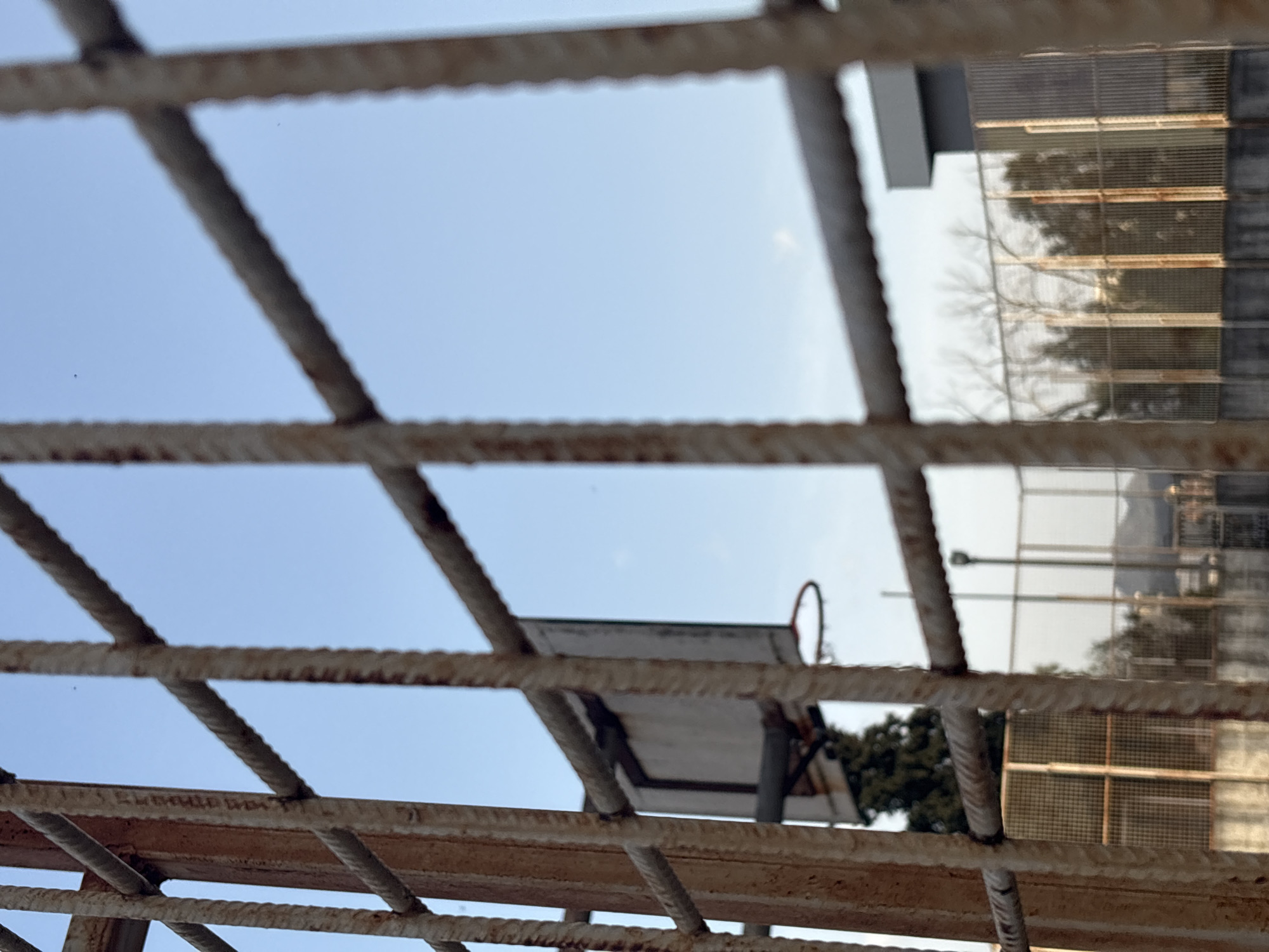 The image is an eye-level, close-up shot of a rusty metal fence in front of a partly visible basketball hoop and a multistory building with a grid pattern facade. The sky is visible through the fence. The scene appears to be in an urban environment. The location may be in Bar, Montenegro, based on the prompt.
