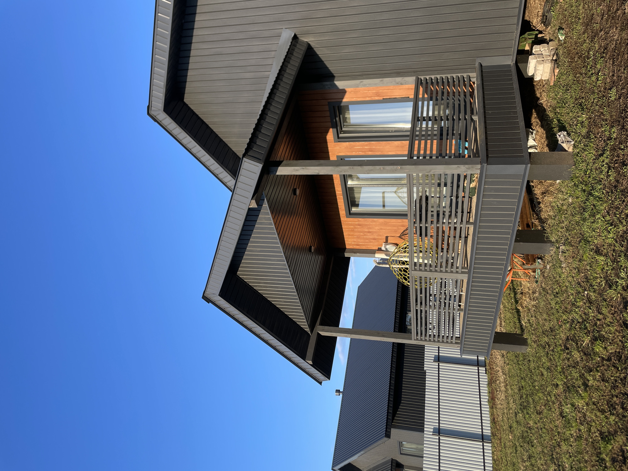 This image captures a modern, two-story house in Podshivalovo, Russia, under a clear, bright blue sky. The house features dark grey corrugated metal siding, complemented by warm wooden paneling on the upper exterior walls around the windows, and on the soffits of the roof and balcony. A prominent balcony extends from the upper level, equipped with dark grey slatted railings. Two windows are visible on this level, reflecting the sky. On the balcony, a distinctive yellow spherical object, possibly a decorative light fixture or part of a chair, is present.
The scene is bathed in bright daylight, as indicated by the deep blue sky and the strong shadows cast by the rooflines and balcony, suggesting it is mid-day or early afternoon. The ground around the house is covered in green and brown grass, appearing somewhat wild or recently developed. Scattered near the base of the house are some concrete blocks and a partially visible orange construction element. In the background, another structure with similar dark corrugated siding is partially visible. No people or visible text are present in the image.