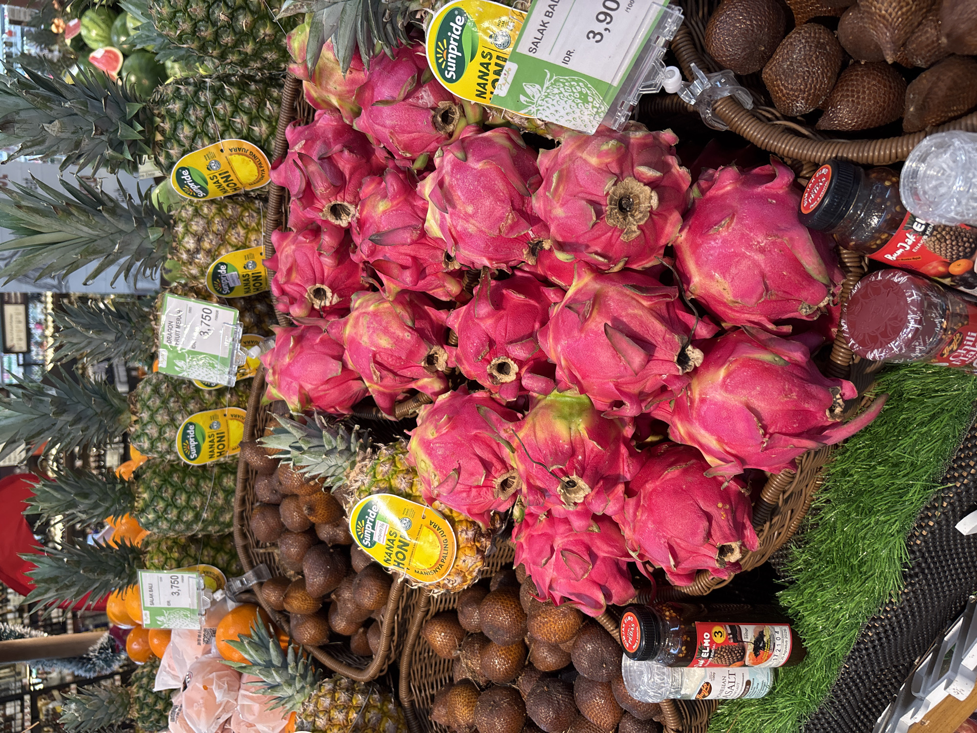 The image shows a fruit stall in Kuta Selatan, Indonesia. The stall is filled with a variety of fresh fruits, including dragon fruits, pineapples, oranges, and salak fruits. The fruits are arranged in woven baskets and on a green artificial grass surface. There are also some bottles of chili sauce and salt on display. The lighting is bright, suggesting an indoor market or a well-lit outdoor area. The overall scene depicts a vibrant and abundant display of fresh produce.