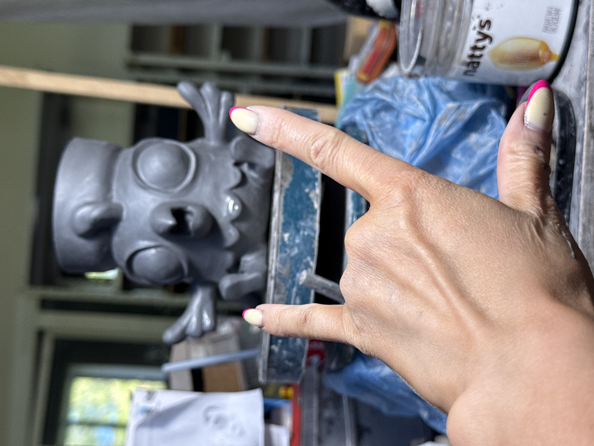 The image shows a person's hand making a "rock on" hand gesture, with the index and pinky fingers extended. The hand is positioned near a grey clay sculpture of a cartoonish head with large eyes and protruding teeth, held in a clamp. Behind the hand and sculpture, there is a jar of "Natty's" brand product and a blue plastic bag. The background includes shelves and a window. There is no information in the image to suggest the location is in the city of Chaikovsky, Russia.