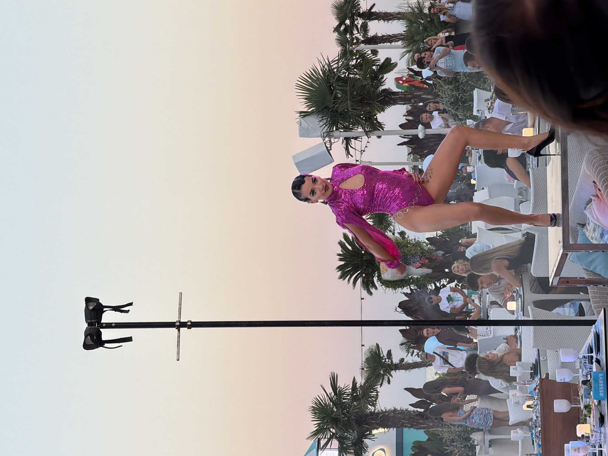 The image depicts an outdoor scene at dusk, possibly at a beach club or restaurant in Odesa, Ukraine, given the presence of palm trees and the general ambiance of a coastal resort. The sky shows a gradient of soft, pastel colors, indicative of sunset or sunrise.
The central figure is a woman performing on a raised platform. She is wearing a bright pink sequined leotard with cutouts and silver chain detailing around the waist and hips. She has dark hair styled in waves and is wearing dark eye makeup. She is also wearing black high-heeled shoes. Her pose suggests a dynamic dance performance.
In the background, there are numerous tables and chairs set up, with diners seated and interacting. Many are dressed casually, reflecting a relaxed evening setting. There are also palm trees and some decorative foliage, contributing to a tropical or Mediterranean atmosphere. A tall pole with what appears to be some sort of lighting or sound equipment is visible behind the performer.
Further in the background, there are more people, some standing and observing the performance, while others are seated at tables. The setting appears to be a popular social gathering spot, likely a restaurant or entertainment venue by the sea. The overall mood is festive and lively, with the performer being the clear focal point of attention.