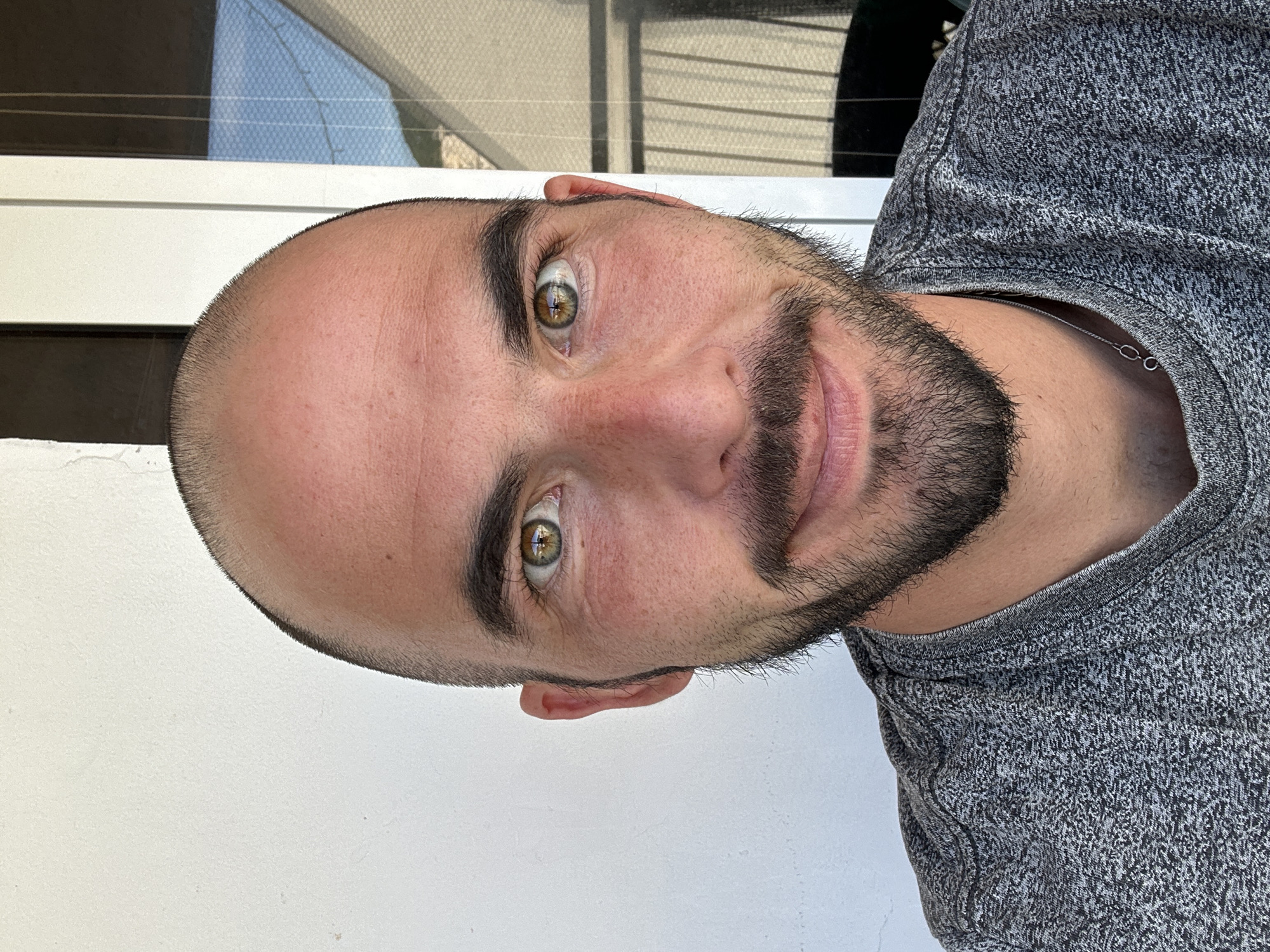 A close-up, head-and-shoulders, eye-level selfie of a man with a shaved head and a goatee. He has brown eyes and is wearing a grey marled t-shirt and a thin silver chain. The man is outdoors, with a white wall on his left and a window with blinds on his right, suggesting a residential setting in Budva, Montenegro. The lighting indicates it is daytime.