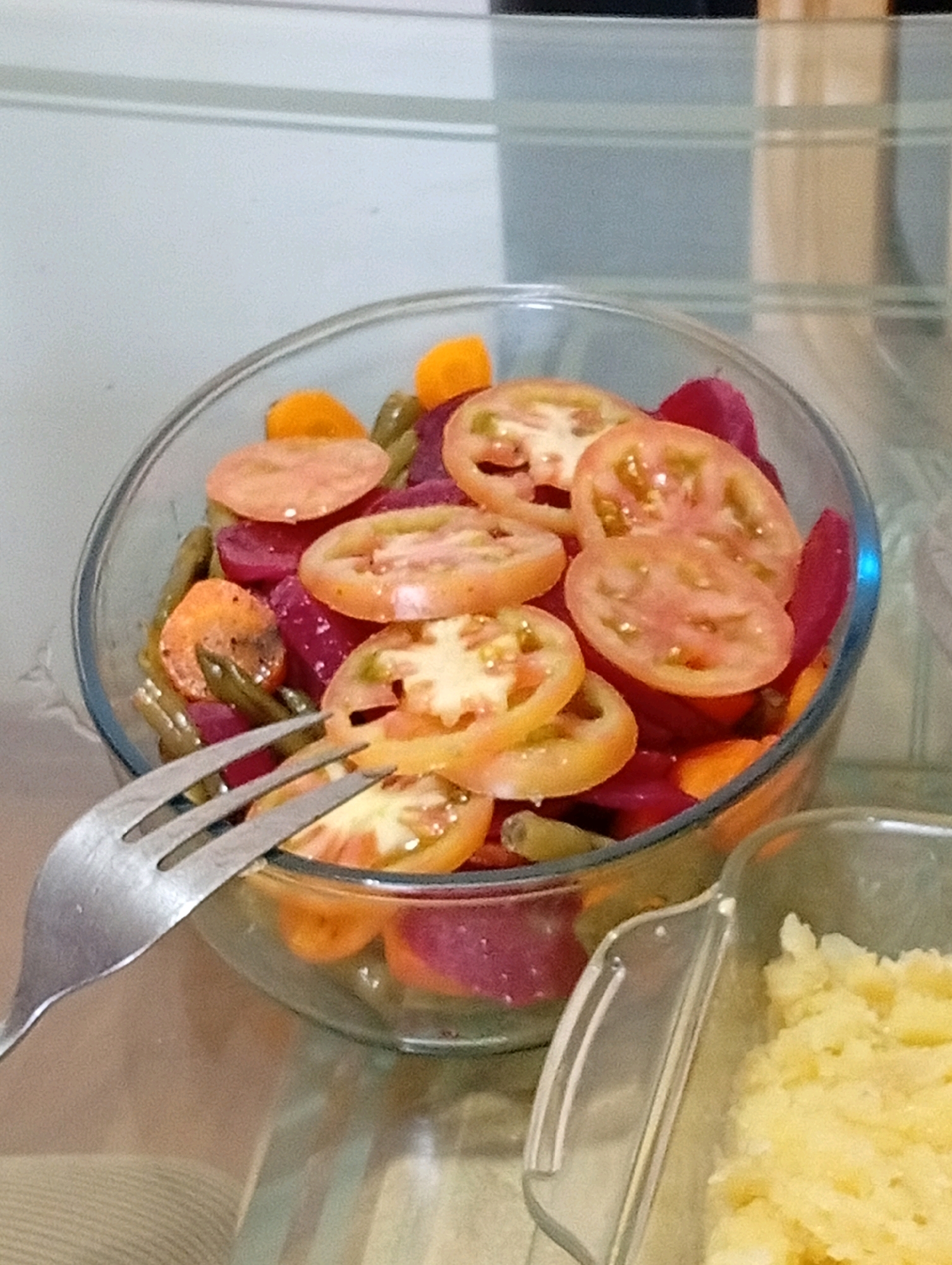 A close-up, eye-level, slightly angled medium shot captures a glass bowl filled with a colorful salad composed of sliced tomatoes, beets, carrots, and green beans. A silver fork rests on the rim of the bowl, its tines dipping into the salad. To the right of the bowl, a clear plastic container holds mashed potatoes. The background is softly blurred, showing a neutral-toned wall with horizontal lines. The lighting is diffused and appears to be indoor ambient light. The scene is set on a reflective surface, likely a table, with the overall impression of a home-prepared meal. The location is identified as San Antonio de Los Altos, Venezuela.