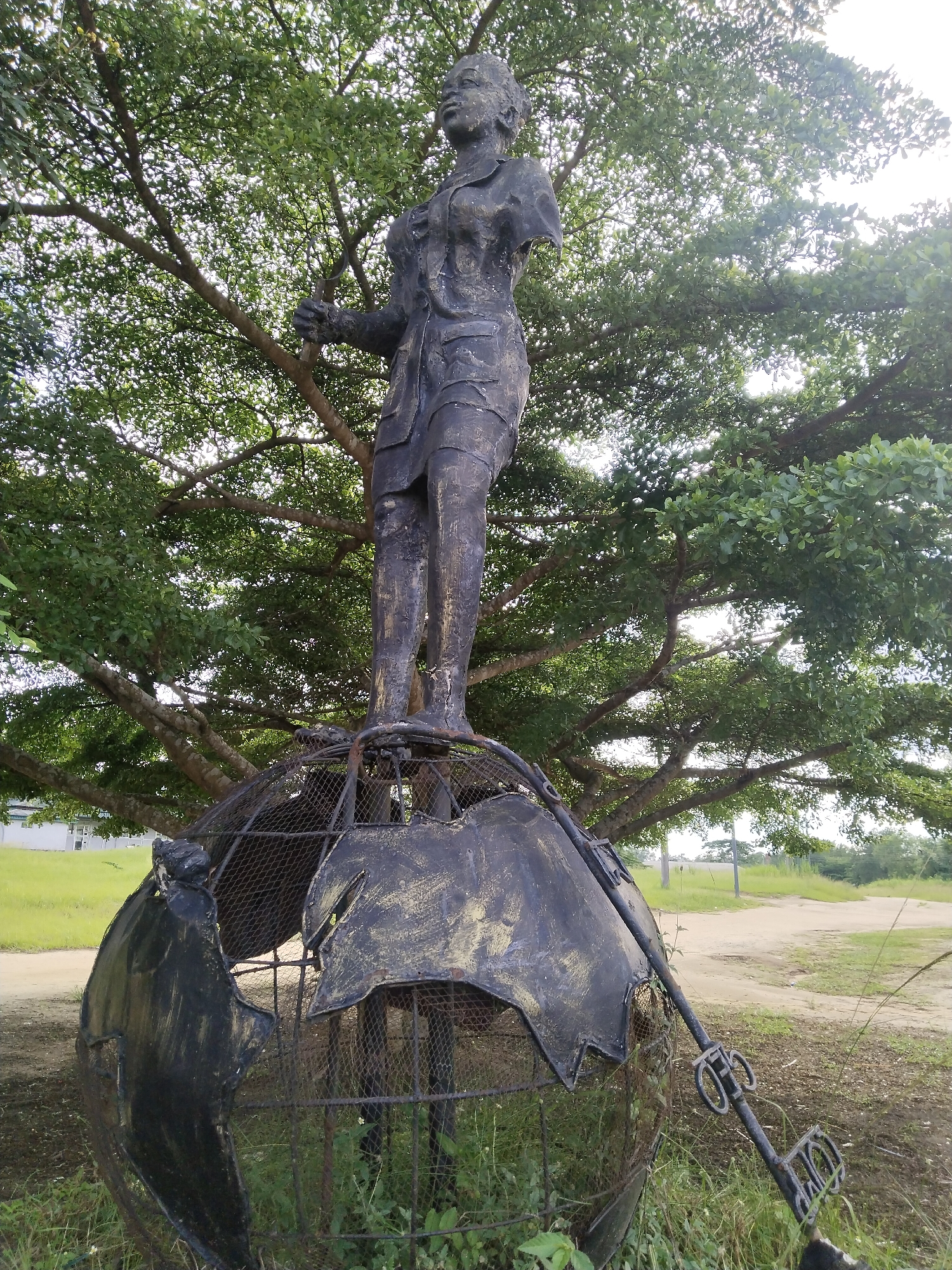 A statue of a woman stands atop a metal globe that is in disrepair. The statue is missing its arms and is weathered, and the globe is also damaged with missing sections and rusted areas. A large tree with dense green foliage looms in the background, partially obscuring the statue and globe. The scene is set outdoors in a grassy area, with a road or path visible in the background. The location is in the city of Uyo, Nigeria.