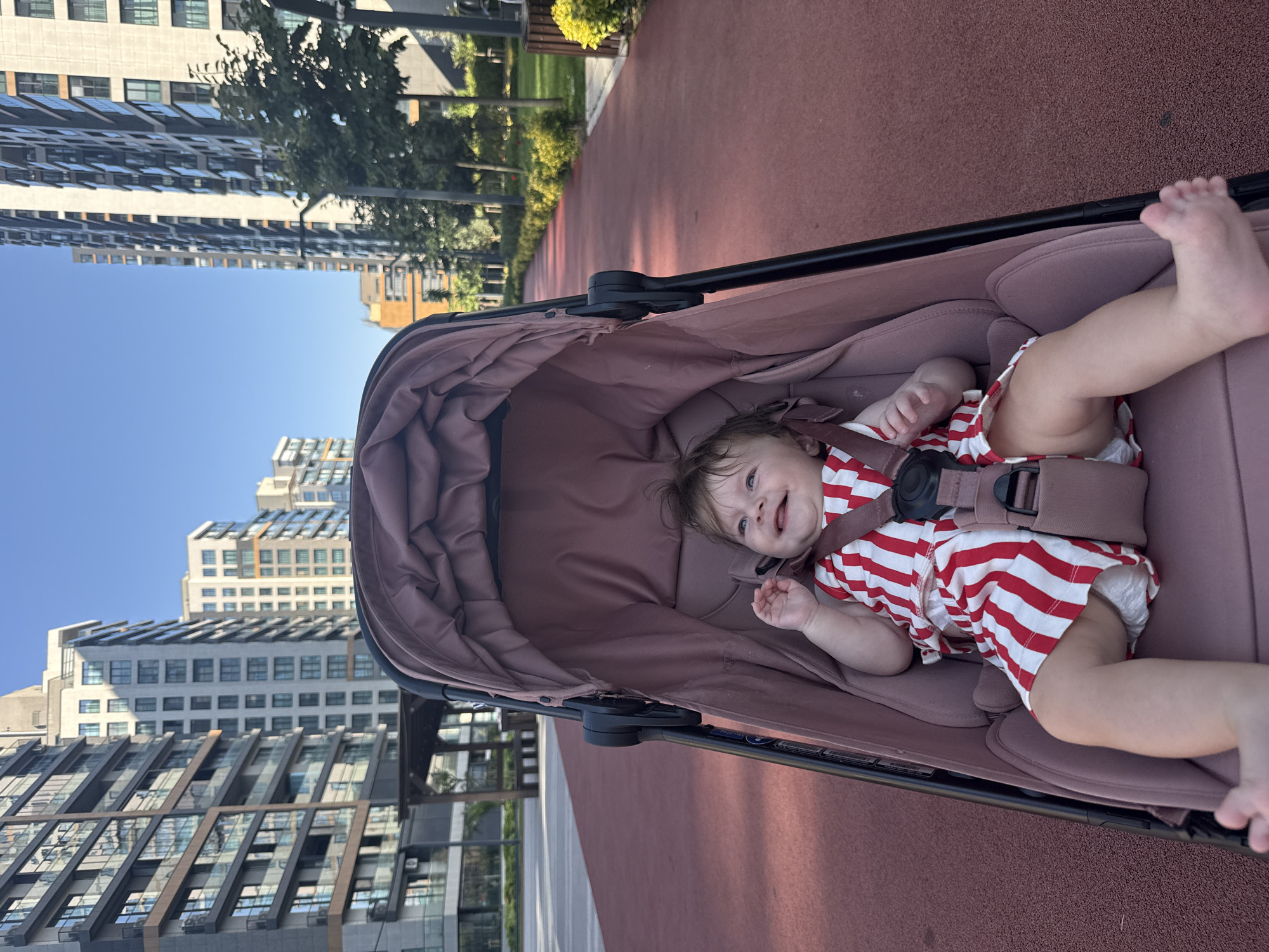 A baby wearing a red and white striped outfit is seated in a pink stroller. The baby is smiling and looking up and to the left. The stroller is positioned on a red pathway in a modern urban environment. In the background, several tall, contemporary buildings with many windows line both sides of the pathway, suggesting a residential or commercial district. Trees and green spaces are visible between the buildings and along the pathway, indicating a planned urban park or communal area. The sky is clear and blue. The location is identified as Ümraniye, Turkey.