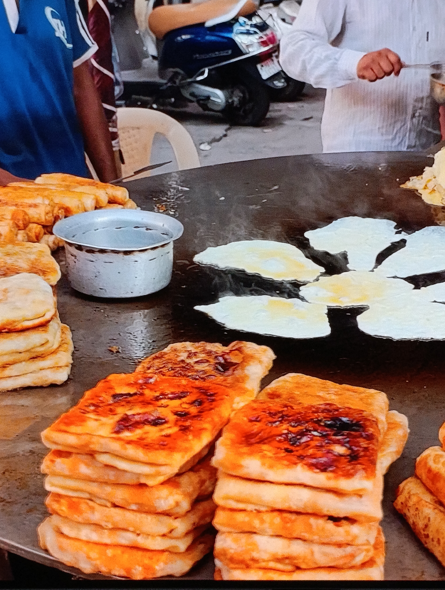 Here's a detailed description of the image:
**Scene:** The image captures a food preparation scene, possibly in a street food setting.
**Subjects & People:**
* Two individuals are partially visible. One person is wearing a blue t-shirt with white accents and the other is in a white long-sleeved shirt and is cooking food on the griddle.
**Objects:**
* A large, round griddle (tawa) dominates the scene.
* On the griddle:
* Several fried eggs are cooking.
* A stack of square, cooked flatbreads with a golden-brown surface.
* Another stack of what appear to be cooked, rolled flatbreads.
* A metal container sits on the griddle.
* In the background, a blue scooter.
**Actions & Interactions:**
* Someone (the person in the white shirt) is cooking food on the griddle.
**Environment:**
* The location appears to be an outdoor setting, potentially a street or market.
* In the background, there's a glimpse of a scooter, indicating an urban environment.
**City and Location:**
* Unknown, Unknown (as specified).