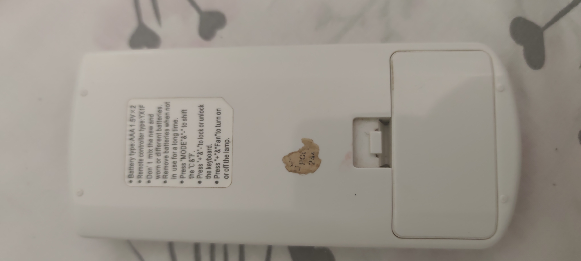 A white remote control is pictured lying on a light-colored, textured surface, possibly a bedspread with a subtle pattern. The remote is oriented horizontally, with the battery compartment cover at the right. Text is visible on the left side of the remote, detailing battery type and usage instructions. A small, worn sticker with faded lettering and numbers is affixed to the lower left of the remote. The image shows the reverse side of the remote. The specific location in Kyiv, Ukraine cannot be determined from the image.