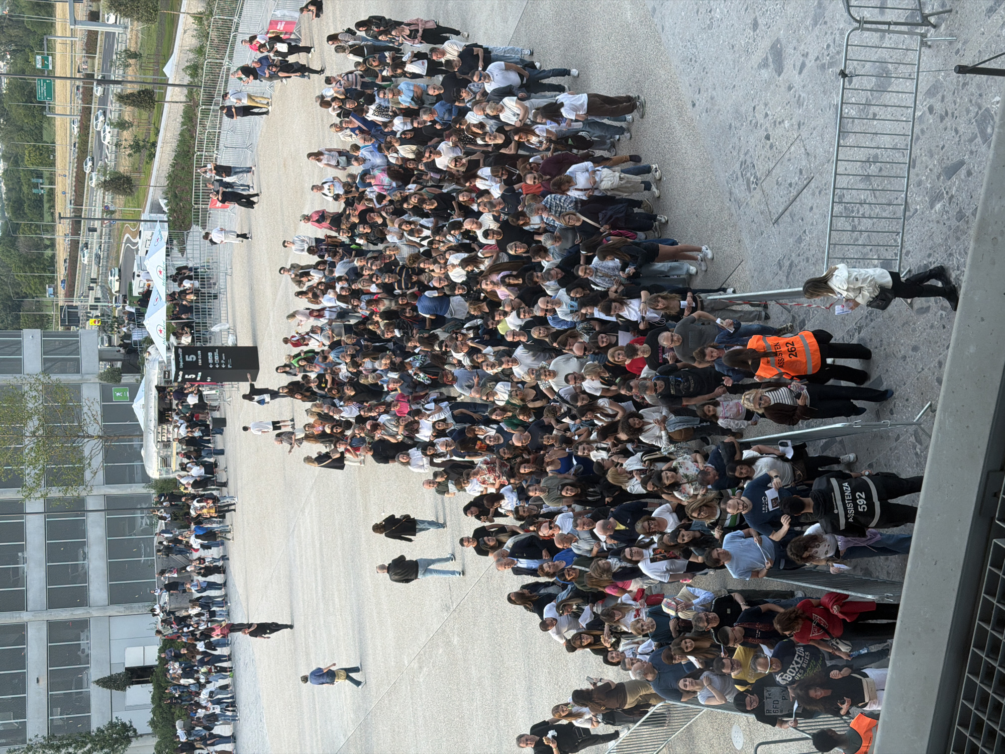 A large crowd of people is gathered in an outdoor area. The crowd appears to be waiting or queuing, as many individuals are facing in the same direction. The setting seems to be in a modern urban environment, with a large building with many windows in the background. Several people stand out, including personnel in orange vests and black uniforms with the word "ASSISTENZA" and the number "592" on the back, suggesting they are security or event staff. Barriers and stanchions are visible, further suggesting a structured queue or controlled access area. A white tent with the Heineken logo can be seen in the background, indicating a sponsorship or event. Based on the style of building and other clues, it seems that this image was taken in the city of Milan, Italy. The exact location is not identifiable from this image alone.