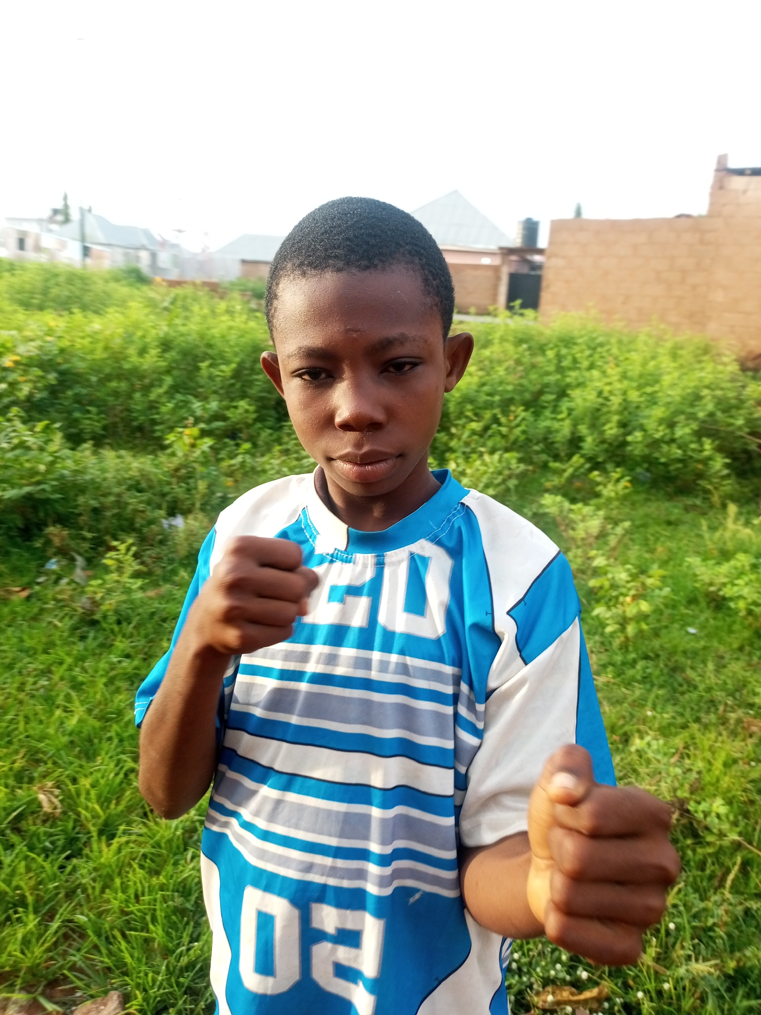 A young Black male stands outdoors in Tempure, Nigeria, facing the camera with a neutral expression. He has short, dark hair and dark eyes. He is wearing a blue and white striped t-shirt with the number "02" on the front. His right fist is clenched and held up in front of him, and his left fist is also clenched and held slightly lower. The background consists of lush green vegetation and some buildings in the distance, suggesting a semi-rural or suburban setting. The sky is overcast.