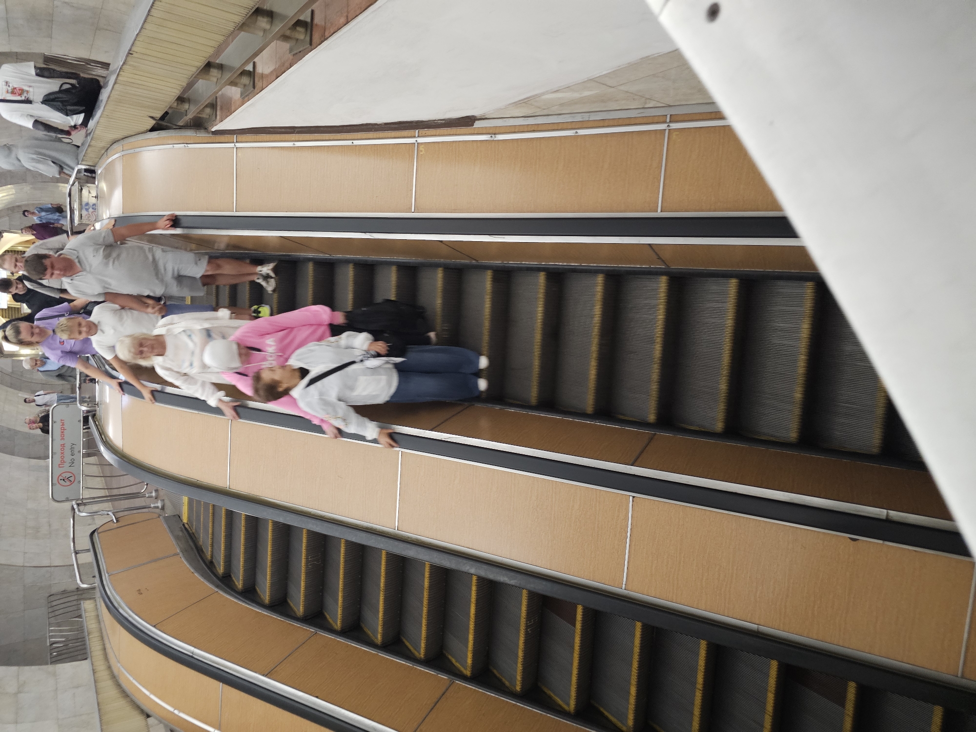 A group of people ride down an escalator in what appears to be a subway station. The escalator is enclosed by wooden-paneled walls and metallic railings. A sign above the escalator reads "Проход закрыт No entry." The people are dressed casually, and some are carrying bags. The lighting in the station is somewhat dim, with overhead lights illuminating the escalators and the surrounding area. The scene captures a common urban commute.