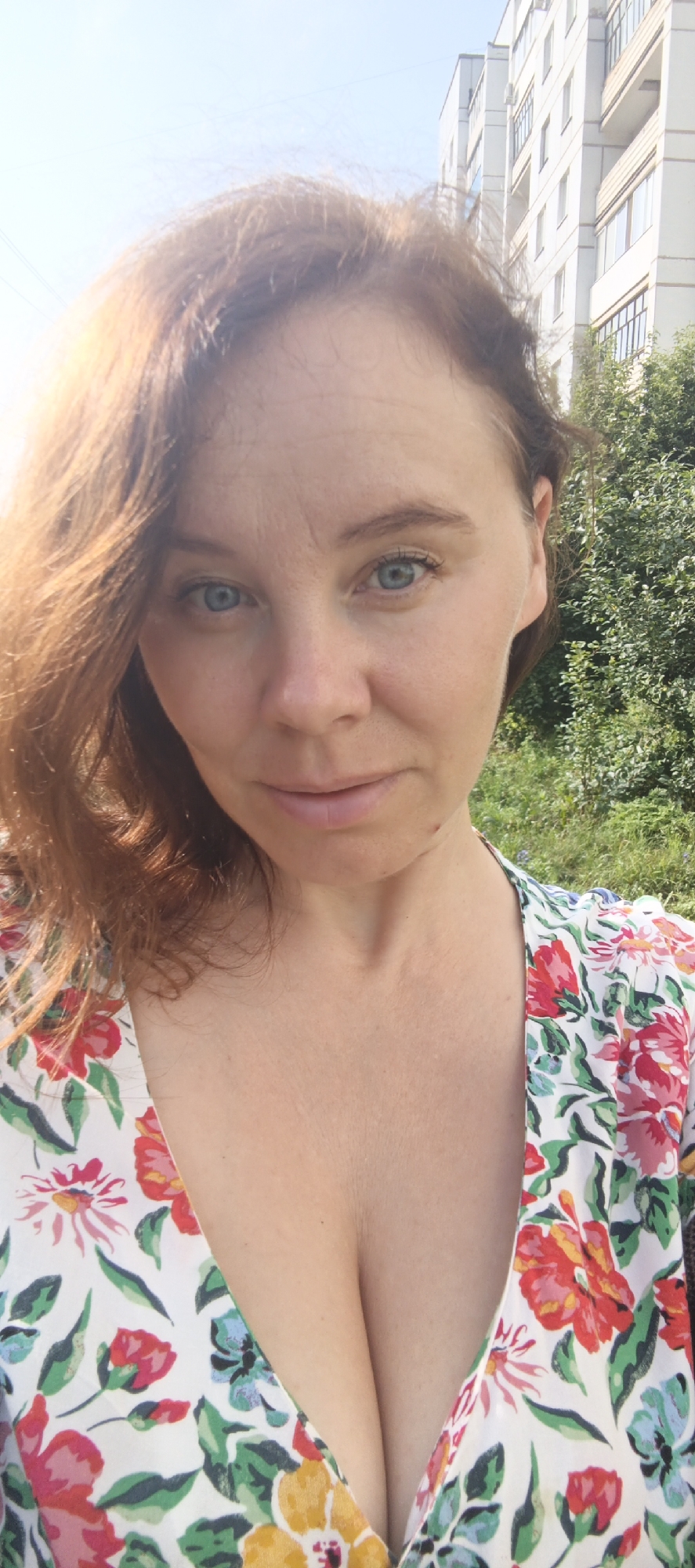 The image is a close-up, outdoor, selfie-style photograph of a woman with light skin and red hair. She is wearing a white dress with a floral pattern featuring red, pink, yellow, and blue flowers, and green leaves. The woman is looking directly at the camera with blue eyes and a slight smile. Her hair is partially obscuring her face due to the angle and wind.
The background shows a bright, sunny sky with a faint indication of power lines. To the right of the woman, there is a multi-story apartment building with white and light gray walls and numerous windows. In front of the building and to the right of the woman, there are green trees and foliage. The overall setting appears to be an urban environment, possibly a residential area, on a clear day. The image is likely taken in the city of Chaikovsky, Russia, as indicated by the architectural style of the building and the surrounding vegetation.
