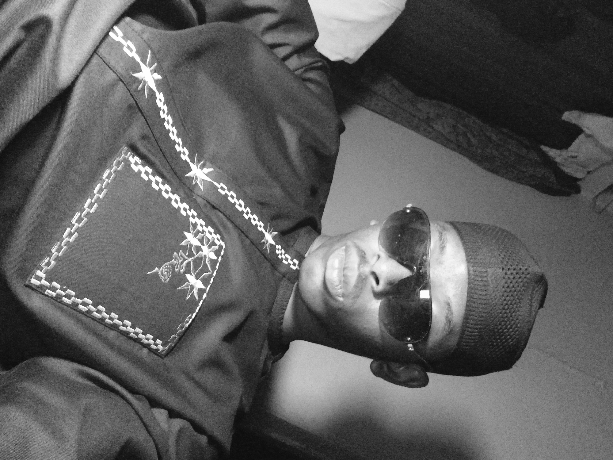 A black and white, angled portrait shows a person wearing a dark shirt adorned with a decorative stitched pattern, a skull cap, and sunglasses. The shirt features a square patch with embroidered details and a decorative stitch pattern running across the chest. The person's face, including the nose and lips, is partially visible behind the sunglasses. The background shows a blurred wall and possibly some bedding. The location cannot be determined, as there is no clear indication of being in Lagos, Nigeria, from the visual details.