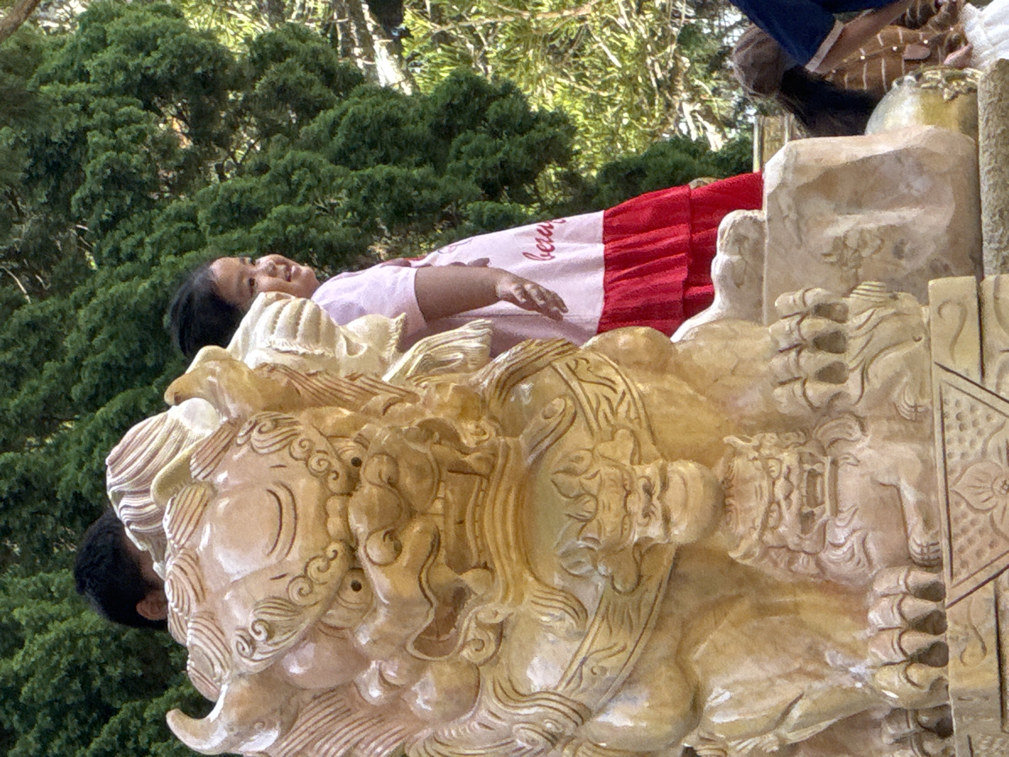 A young girl in a pink and red dress stands to the right of a large, light-colored stone statue of a Foo dog. Her head is tilted slightly upwards and to the left, and she is smiling. The Foo dog statue is intricately carved with detailed facial features, mane, and claws. Behind the girl and to the left of the statue, the back of a person's head is visible. The background consists of lush green trees. The scene appears to be outdoors, possibly in a garden or park. The presence of the Foo dog statue suggests a possible location in an Asian cultural setting, and the style of the statue is consistent with architectural elements found in Da Lat, Vietnam, known for its French colonial and Asian architectural influences.