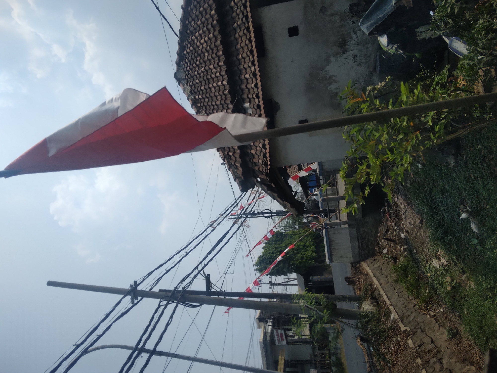 The image captures a street scene in Wringinanom, Indonesia, under a partly cloudy sky. In the foreground, an Indonesian flag, predominantly red and white, is prominently displayed on a wooden pole, angled towards the viewer's right. The flag appears slightly blurred, suggesting movement. Behind the flag, a simple, weathered building with a tiled roof is visible. A network of utility wires, supported by poles, spans across the upper portion of the frame, indicating an urban or semi-urban setting.
To the left of the building and flag, a road runs horizontally, with a concrete channel or small canal bordering the foreground. Along the edge of this channel, there is a mix of vegetation, debris, and a stone retaining wall. Small Indonesian flags on poles are sparsely distributed along the roadside and in the distance, adding a festive or patriotic element to the scene. In the background, other buildings and trees are visible, suggesting a residential or commercial area. There are no people clearly identifiable in the immediate foreground or midground, though the presence of a motorcycle parked near the building in the right midground suggests human activity. The overall impression is one of a typical day in a developing area, possibly decorated for a national celebration.