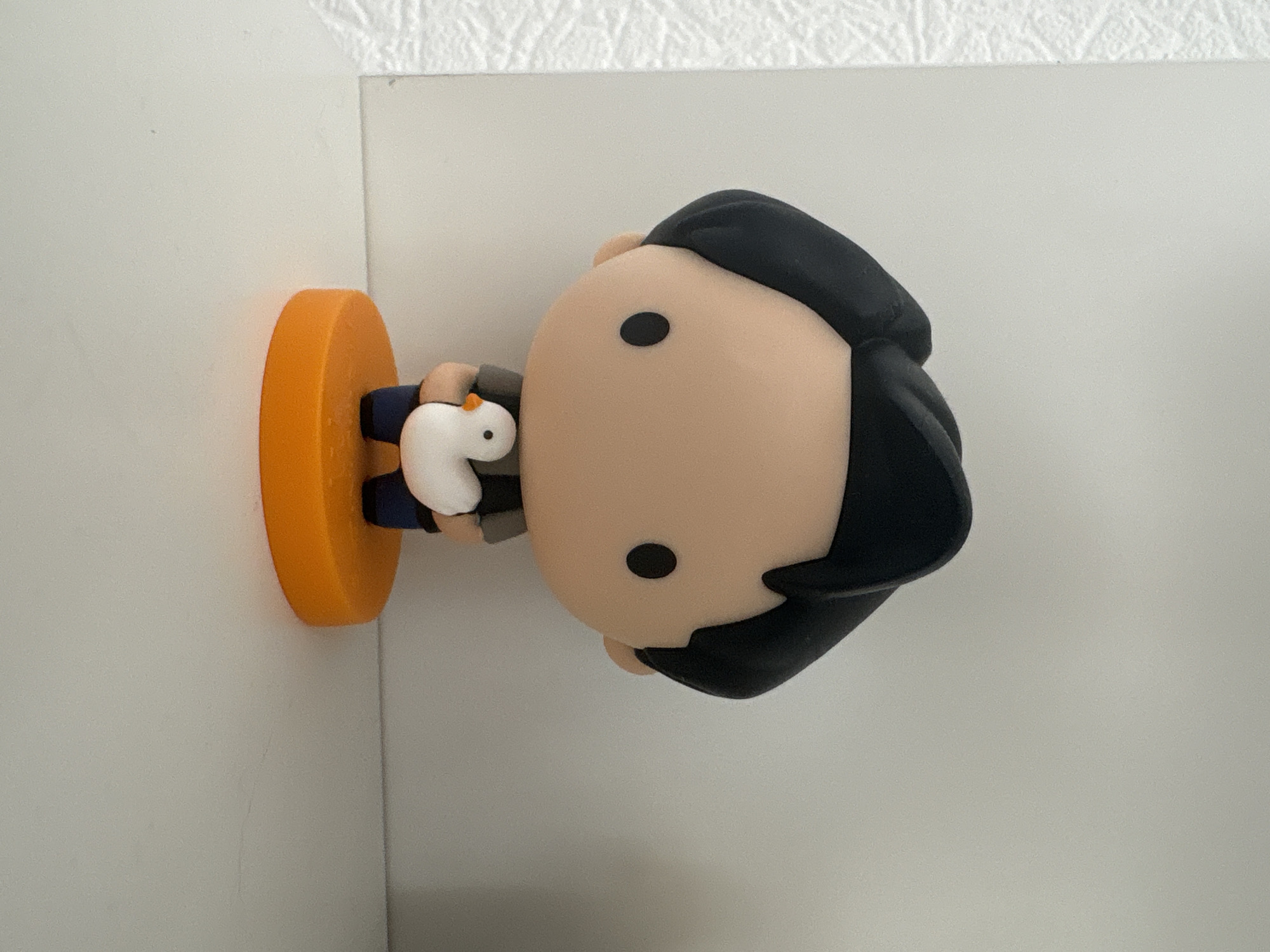 A vinyl toy figure of Chandler Bing from the TV show *Friends* is positioned on a white shelf against a white background. The figure is approximately six inches tall, featuring a black hairstyle, a pale face with black dots for eyes, and is holding a small white duck. The toy stands on a bright orange circular base. The environment appears to be an indoor setting, potentially a home or an office, located in Kyiv, Ukraine.