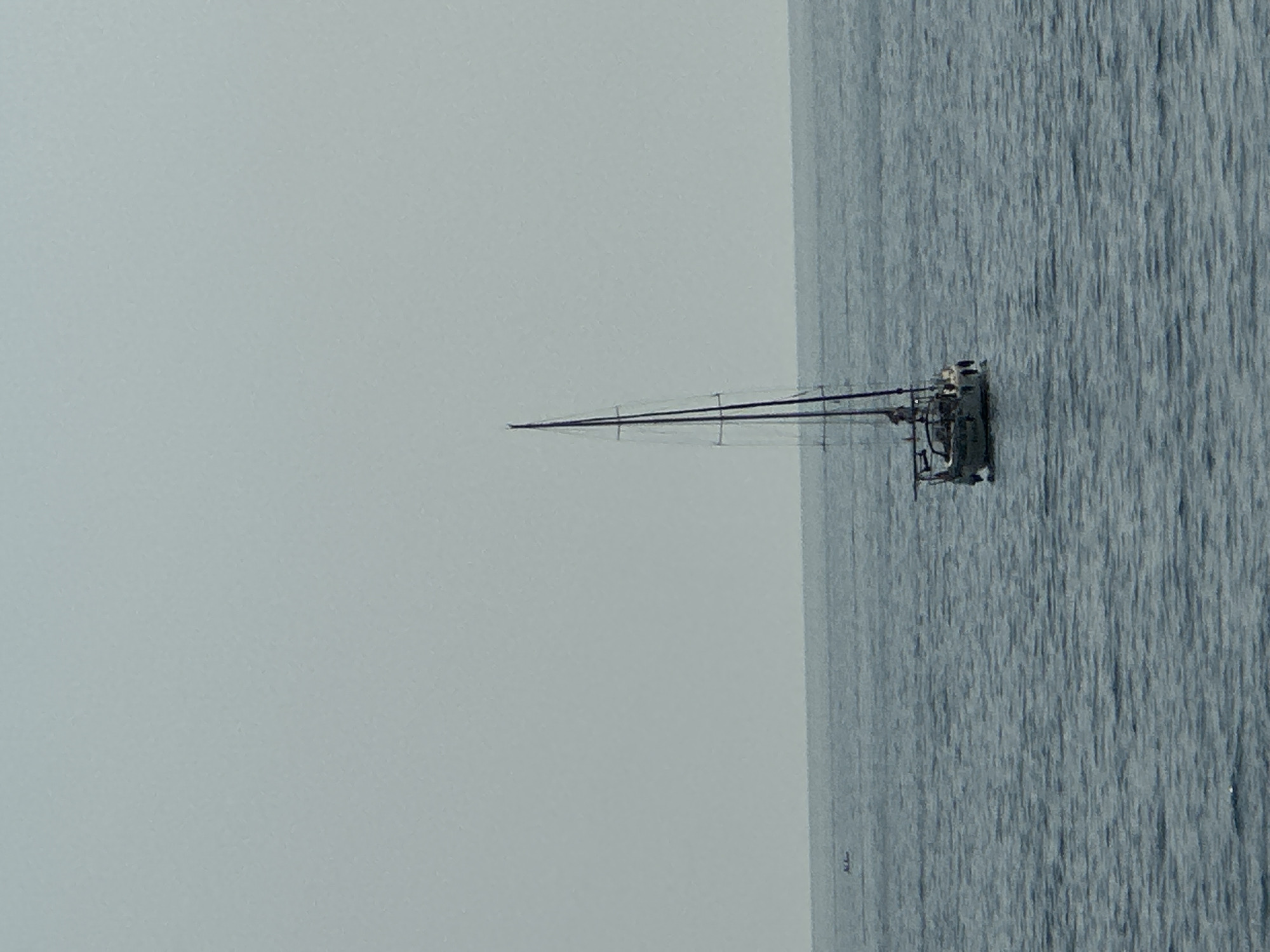 A sailboat is visible in the open sea, under a hazy, pale sky. The boat is white with a dark mast, and there appear to be people on board. In the distance, a smaller, dark vessel is also visible on the water. The sea is a muted blue-grey, with gentle waves. The horizon is a distinct line separating the sea from the sky. The scene is calm and expansive.