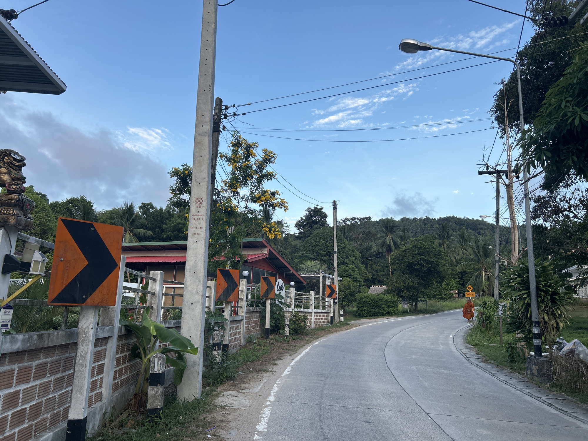 This image depicts a winding paved road in the Ko Pha-ngan District of Thailand, with lush green vegetation and trees lining both sides. The sky is a vibrant blue with scattered white clouds. On the left side of the road, a brick wall runs alongside, with traffic signs indicating sharp curves. A concrete utility pole stands prominently, supporting electrical wires and a street lamp. A building with a red roof and a light-colored exterior is visible behind the wall. Further down the road, a figure in an orange robe, likely a monk, is walking away from the viewer. Additional utility poles and dense foliage contribute to the tropical ambiance of the scene.
