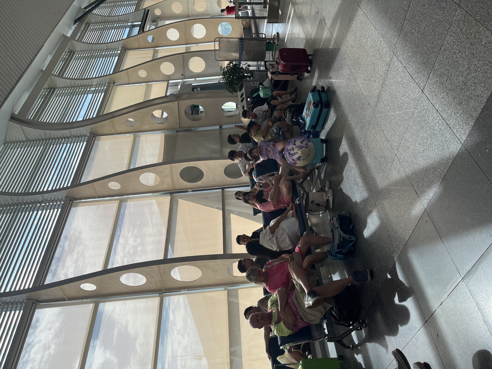 The image displays a brightly lit indoor public waiting area, identified as being in Đà Nẵng, Vietnam, likely an airport or major transit terminal. The space is characterized by a high ceiling and large, multi-paneled windows that span the height of the wall. The upper sections of the windows are fitted with horizontal blinds, while the lower sections allow sunlight to stream in, illuminating the scene and casting prominent shadows on the light grey tiled floor. Structurally, the windows are segmented by prominent, light-colored, curved panels featuring evenly spaced circular cutouts, which serve as both support and architectural detail.
Approximately 15 to 20 individuals, comprising adults and children, are seated across several rows of interconnected blue public waiting chairs. In the foreground, a bald man with a beard, wearing a green and pink patterned shirt and khaki shorts, sits with his arms crossed, next to a blonde woman in a pink t-shirt and shorts. Other groups, predominantly appearing to be East Asian, are also seated, some engaged in conversation, one adult holding a child.
Numerous pieces of luggage are distributed on the floor around the seated individuals. These include a bright green carry-on suitcase on the far left, various backpacks and duffel bags, a light blue hardshell suitcase topped with a decorative circular item, another light blue hardshell suitcase, a beige hardshell suitcase, and a prominent red hardshell suitcase in the right foreground. A potted plant is situated near a metal information stand or barrier in the middle ground, right of center. The overall impression is one of travelers waiting in a modern public facility during daylight hours.