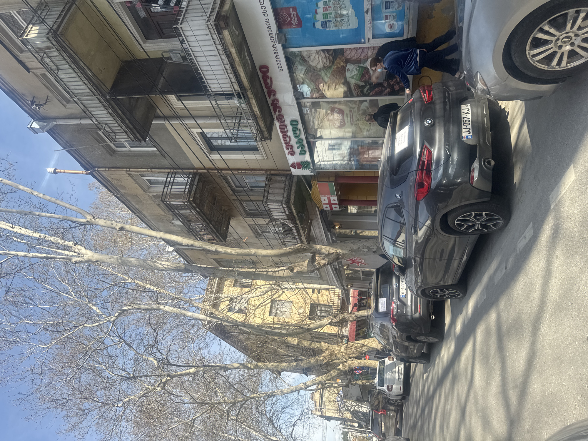 The image captures a street scene in Tbilisi, Georgia. A row of parked cars lines the right side of the street, including a dark-colored BMW with the license plate "JJ-057-KJ" and a dark SUV with the license plate "YC-422-YC." A silver car is parked in front of the BMW, and other vehicles are parked down the street.
On the left side of the street, a building with balconies faces the viewer. A commercial establishment labeled "სახლი ეტების ქსელ" ("Home Network") is situated on the ground floor. "Супермаркет" ("Supermarket") is written in red lettering vertically on the building's facade. A person wearing a blue jacket and a face mask is standing near the entrance.
Bare trees are visible along the street, adding an element of seasonality to the scene. The sky is clear and blue, indicating fair weather.