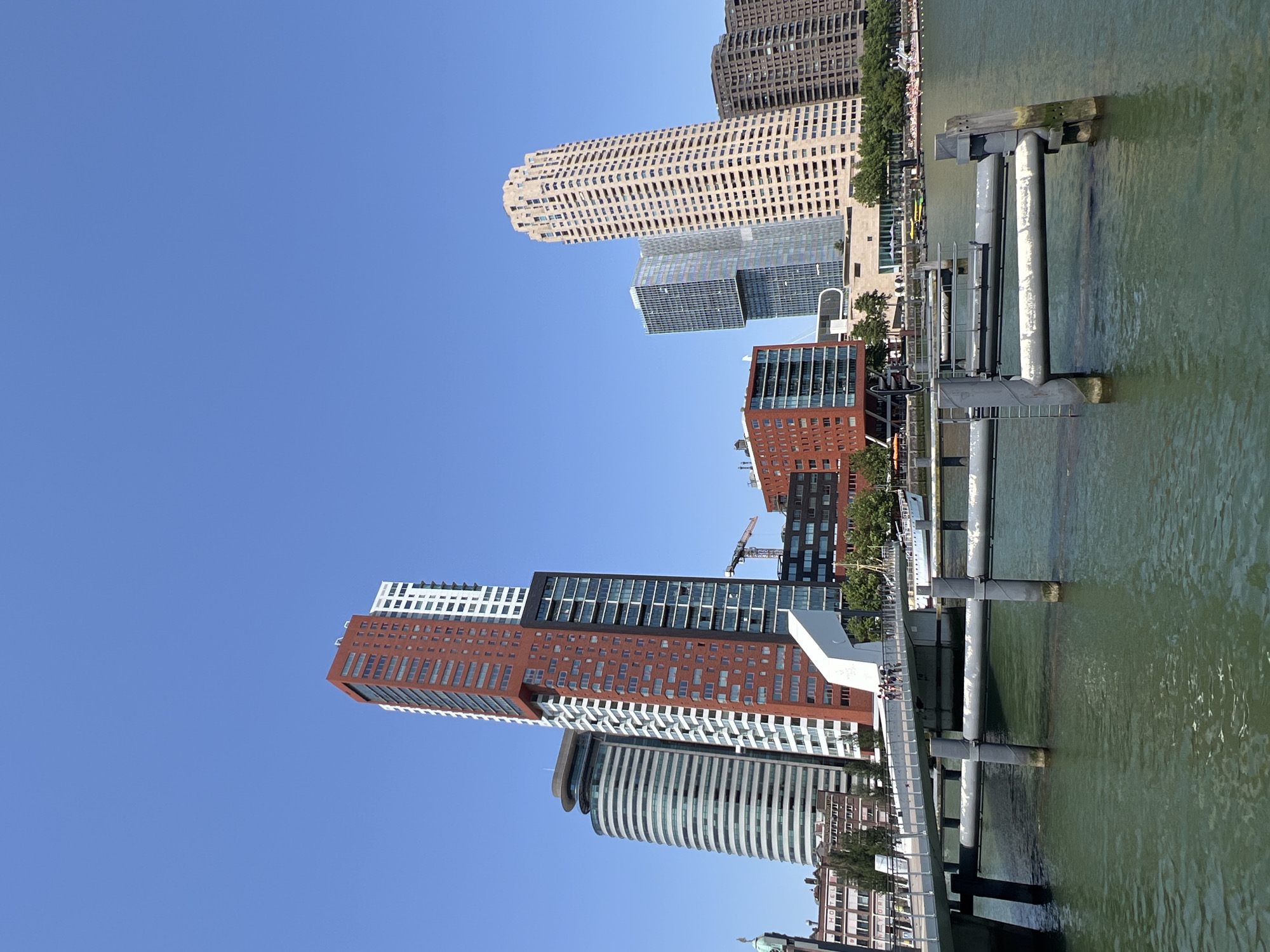 The image depicts a modern cityscape of Rotterdam, Netherlands, characterized by a collection of contemporary skyscrapers and a waterfront promenade. The buildings showcase a variety of architectural styles, including a prominent red and white tower, a rounded, striped building, and a tall, beige, textured skyscraper. In the foreground, a metallic pedestrian bridge extends over a body of green water, supported by dark pilings. Several people are visible walking on the bridge. The water reflects the clear blue sky, which dominates the upper portion of the image. The scene is bathed in bright sunlight, suggesting a clear day.
