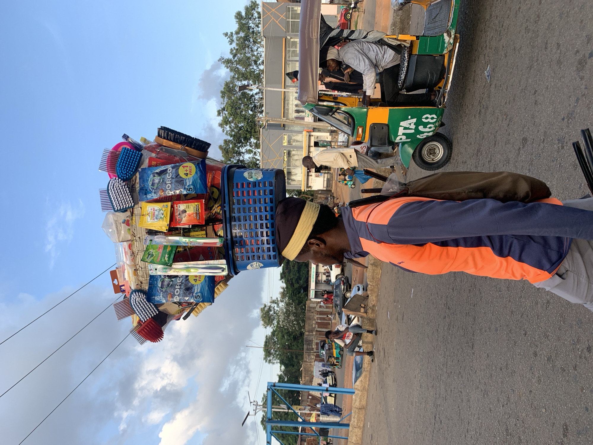 A man wearing a yellow and brown skullcap and an orange and dark blue long-sleeved shirt walks past a street scene in Jos, Nigeria. He carries a large assortment of personal care items, including toothbrushes and brushes, balanced precariously on his head. The items are displayed in blue plastic baskets and are predominantly packaged in plastic and cardboard.
In the background, a green and yellow auto-rickshaw, commonly known as a 'Keke Napep,' is parked on the right side of the frame. A man in a patterned shirt is visible in the driver's seat, and another man in a dark jacket is standing beside the vehicle. Further in the background, there are several buildings, including what appears to be a bus stop with a sign reading "Old Airport Bus Stop," and a row of shops with gray and patterned facades. Other people are seen walking and sitting on a low wall in the middle ground. A blue metal structure, possibly part of a street stall or market, is visible on the left. The sky is partly cloudy, indicating daytime.