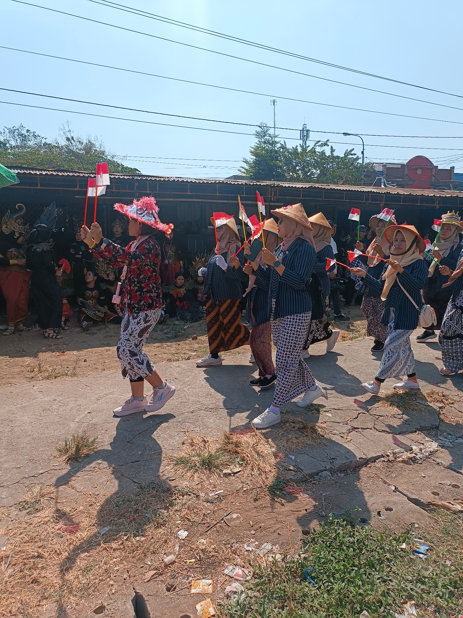 In Imogiri, Indonesia, on a sunny day, a group of women dressed in traditional attire participates in a procession. The women wear straw hats and outfits featuring striped tops and patterned trousers. Some of them are holding small Indonesian flags. They are walking on a paved road with debris and dry grass on the sides, casting long shadows. In the background, there are buildings with corrugated roofs and trees. Other participants in traditional costumes, some wearing black and red attire, are visible in the background, observing the procession. The scene appears to be part of a cultural event or celebration.