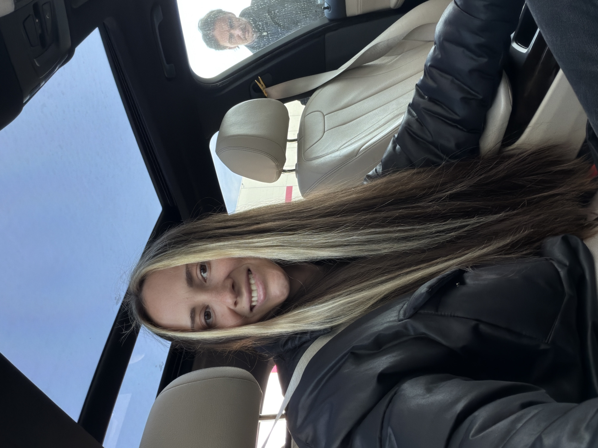 A woman with blonde highlights is seated in a car with a sunroof. She is smiling and looking at the camera. She is wearing a black jacket. The interior of the car is light-colored. Through the sunroof, a man is looking down into the car. The location appears to be outdoors during daylight, with a visible blue sky. The setting of Sluchainyi, Russia is not ascertainable from the image.