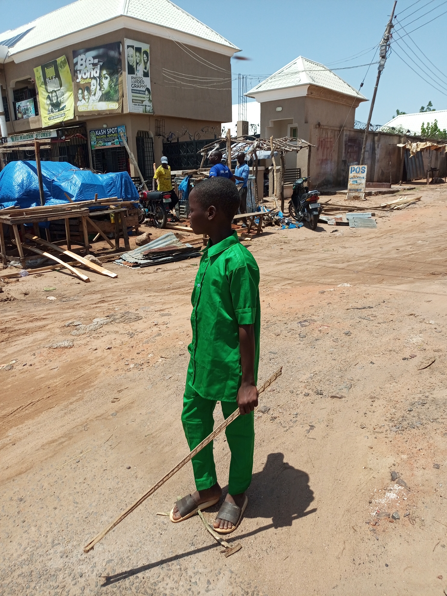 This media file shows an outdoor scene on a sunny day, likely in a developing country given the unpaved road and buildings. A young boy, dressed in a bright green uniform, stands in the foreground, holding a long stick. He is looking towards the right of the frame. In the background, there are several buildings. One building on the left has large banners advertising "Beyond Studio" and "PHOTO & VIDEO GRAPHY," along with contact information. Next to it, a shop labeled "KASH SPOT POS BUZINESS VENTURES" is visible. Further in the background, another building with a distinctive pointed roof can be seen, and a "POS SERVICE" sign is posted on a wall. Various objects are scattered around the scene, including construction materials like wooden planks and corrugated metal sheets, as well as a motorcycle. The ground is dusty and uneven, with some scattered debris. The overall atmosphere appears to be one of everyday life in a busy, possibly commercial, area.