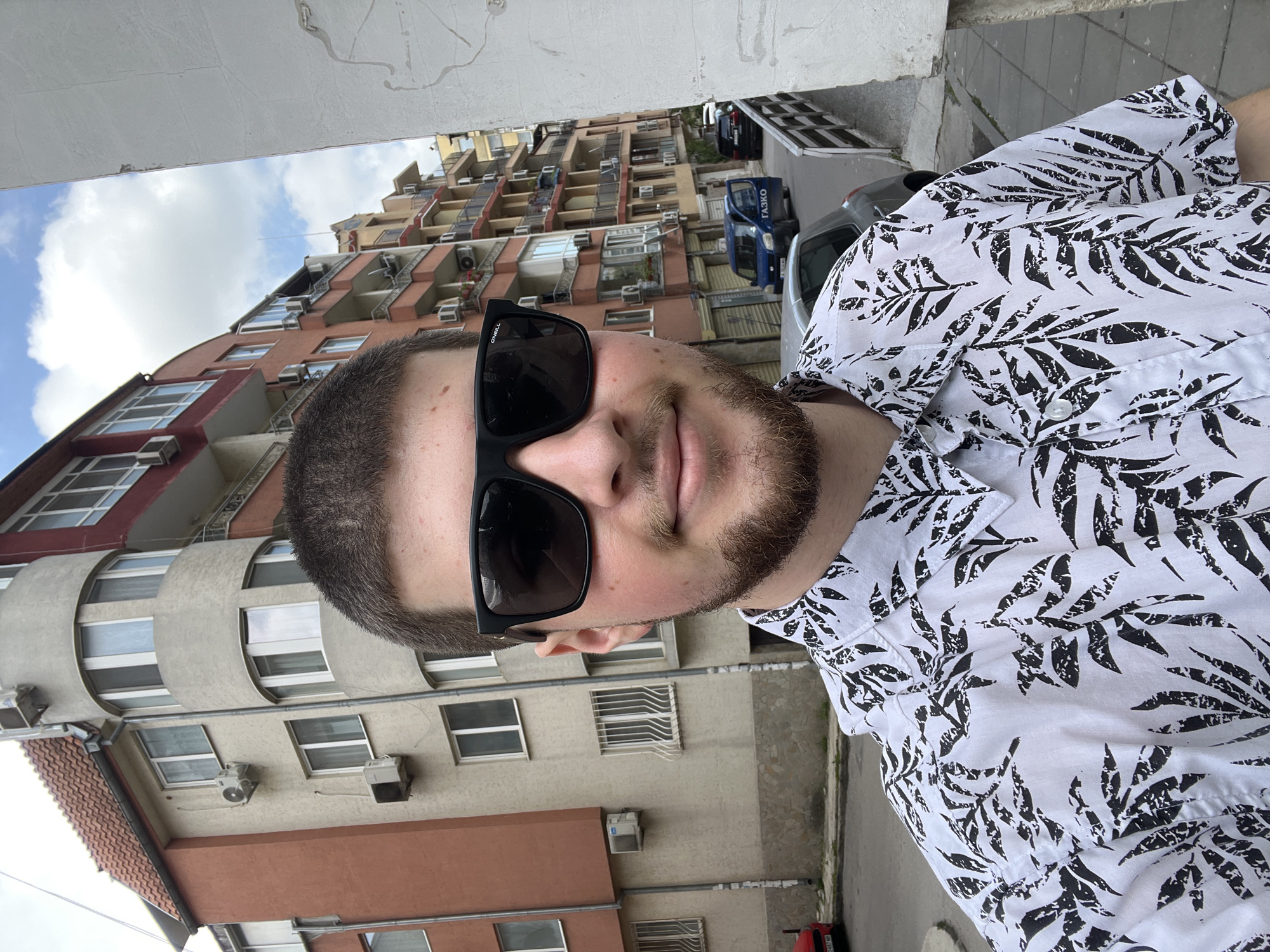 A selfie shows a man wearing a white shirt with a black leaf pattern and black sunglasses, standing outside. The buildings in the background appear to be residential apartments, some with air conditioning units. Parked vehicles are visible further in the background. The location is likely Varna, Bulgaria, based on the architectural style of the buildings and the appearance of a "ГАЗКО" van.