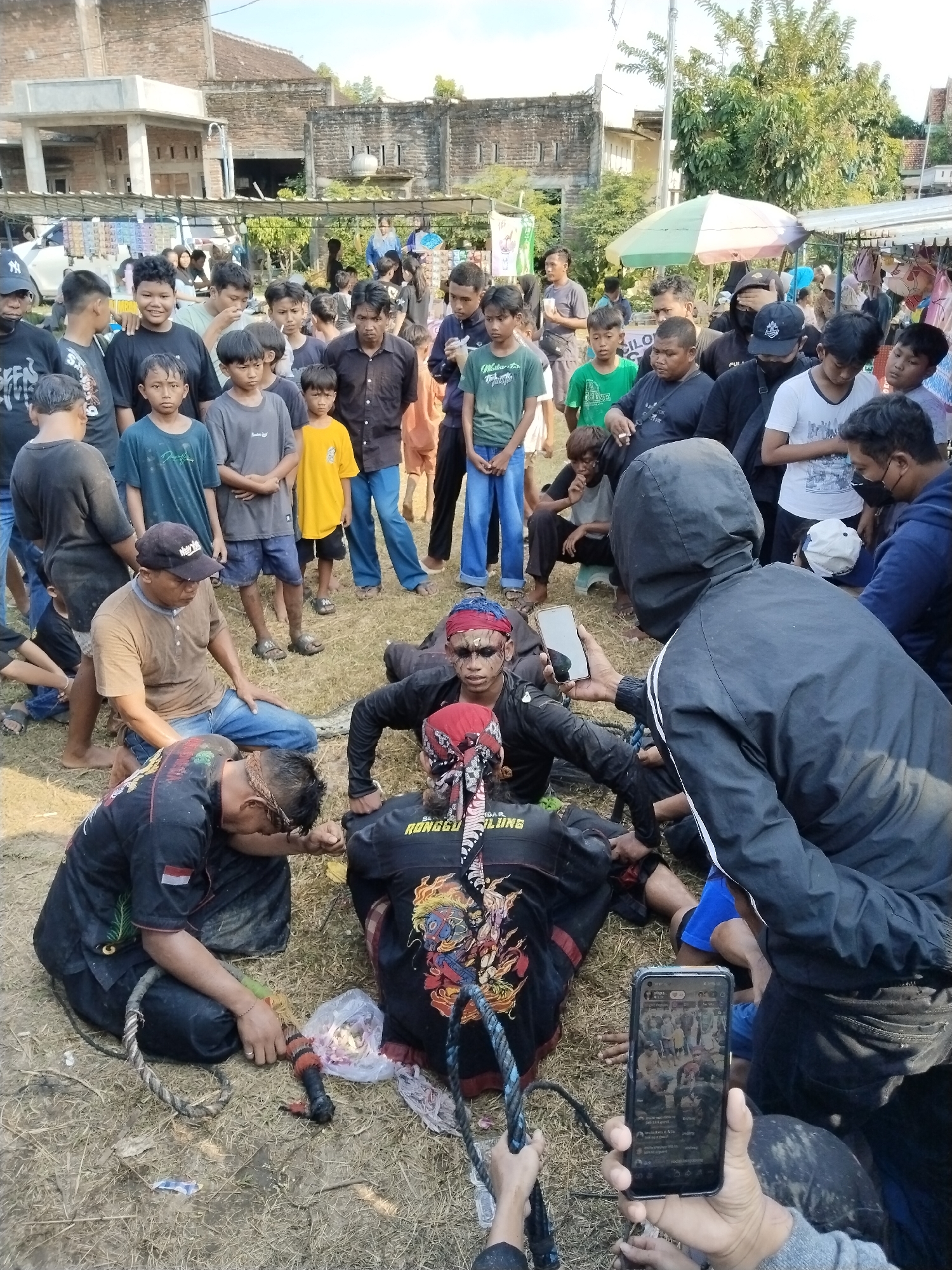 This image captures a vibrant community gathering in Kedawung, Indonesia, centered around what appears to be a cultural or ritualistic performance. The scene is outdoors on dry, dusty earth under bright daylight, likely in the late morning or afternoon.
The primary subjects are several men, some dressed in traditional performance attire. Two men in black shirts with colorful graphics on their backs (one clearly showing "RONGGO SADAR NGELUNG") wear red headscarves and striking dark face paint around their eyes and noses. They are seated or lying on the ground, seemingly in a state of rest or trance, with coiled ropes or whips nearby. One has a small Indonesian flag patch on his sleeve. A man in a brown polo shirt and blue jeans kneels beside them, his hand gently on one of the performers.
Surrounding this central group is a crowd of onlookers, predominantly men and boys of various ages, observing intently. Some children are seen smiling or looking with curiosity. The atmosphere suggests a public event, with many individuals holding smartphones, documenting the occasion. One person in a black hooded jacket is seen holding a smartphone up, possibly recording, while another hand in the foreground holds a phone displaying a social media feed, likely TikTok, showing multiple images from the event.
In the background, simple brick buildings, some appearing aged or unfinished, line the perimeter, along with trees and a striped umbrella. The ground is dry, indicating either a dry season or an unpaved open area. The overall impression is that of a local cultural event, perhaps a traditional dance or trance ritual, drawing significant community engagement and modern-day documentation.