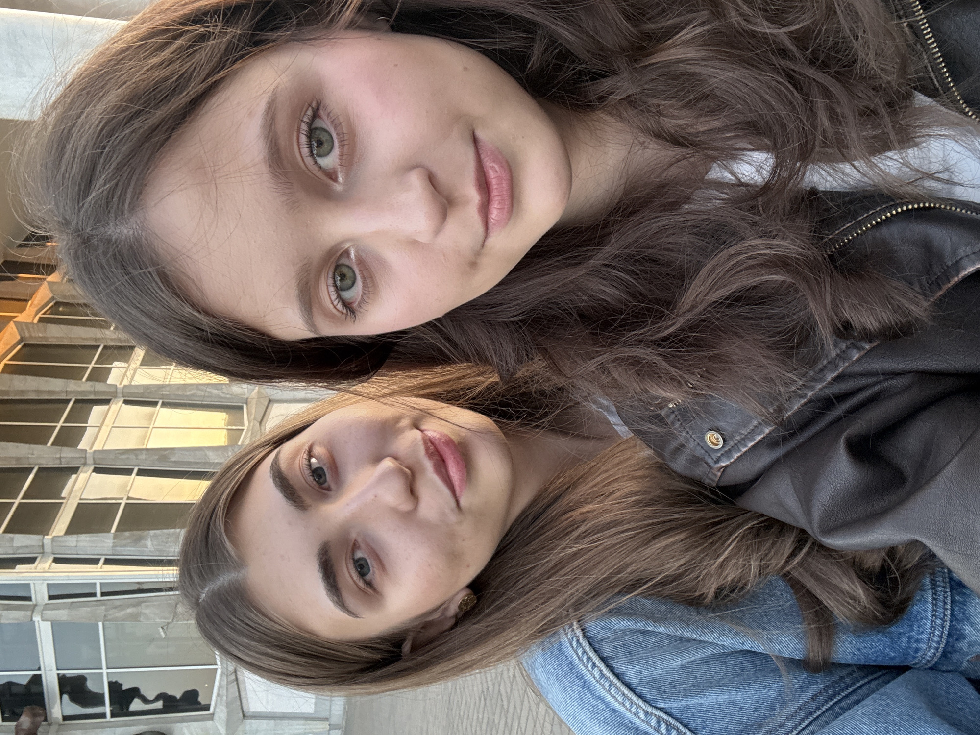 Two young women are posing for a selfie. The woman at the top has light eyes and brown, wavy hair and is wearing a black leather jacket. The woman at the bottom has light eyes and long, straight brown hair, wearing a denim jacket. They are close together. In the background is a light-colored building. The image appears to be taken outdoors, possibly in the city of Krasnoyarsk, Russia. No specific action or interaction is readily apparent aside from posing for the photograph.
