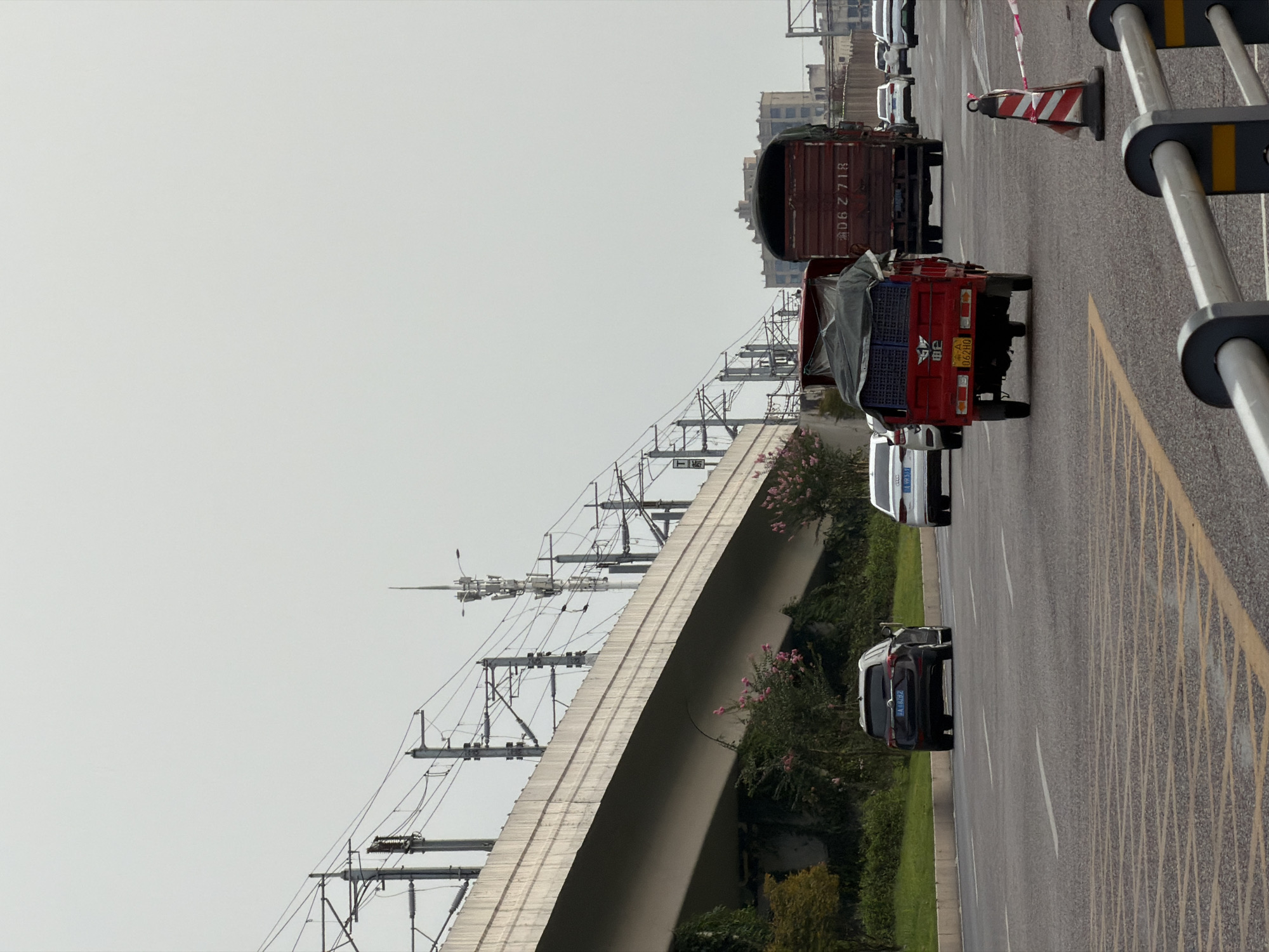 The image depicts a street scene in Jiangjin, China, under an overcast sky. An elevated highway structure with overhead power lines dominates the upper portion of the frame. On the street below, a red three-wheeled truck is in motion, carrying blue crates under a gray tarp. To its right, a large brown truck with the license plate "D6Z 718" is also moving. Several other vehicles, including white cars and a dark-colored SUV with the license plate "A T6282", are visible on the road. Green vegetation with pink flowers lines the side of the road, and buildings can be seen in the background. A traffic cone with red and white stripes is placed on the right side of the road.