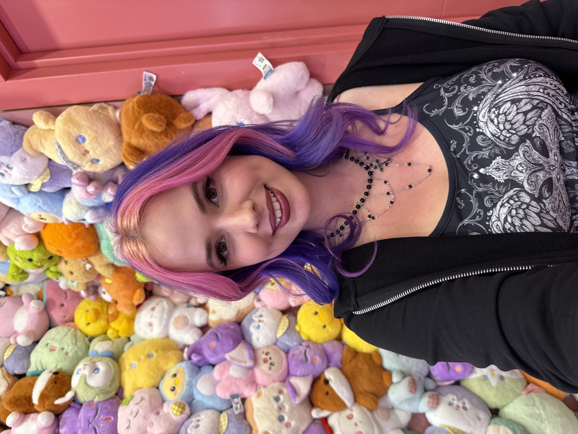 A woman with light skin, pink and purple hair, and wearing a black shirt and jacket lies on a bed of stuffed animals. She is smiling and looking at the camera. The stuffed animals are in a variety of colors and shapes. A red wall is visible in the upper left corner. There is no evidence of the city of Madrid, Spain, in the image.