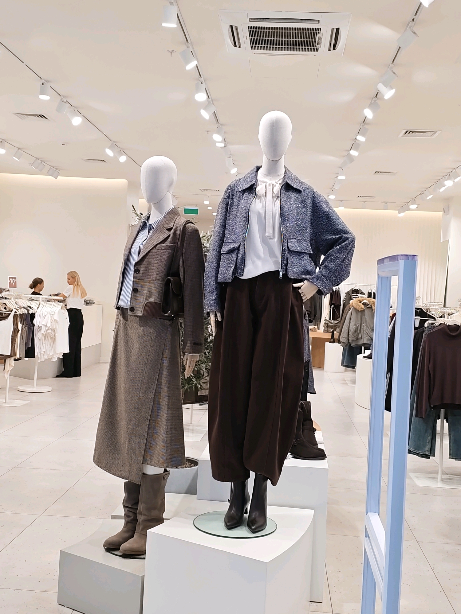 In a brightly lit clothing store in Rostov-on-Don, Russia, two mannequins are prominently displayed. The mannequin on the left is dressed in a brown plaid jacket and a matching skirt, paired with a striped shirt and knee-high grey boots. A dark handbag is slung over its shoulder. The mannequin on the right wears a textured blue jacket over a white blouse, complemented by wide-leg brown trousers and black ankle boots.
Behind the mannequins, a rack of white t-shirts is visible, with two women browsing nearby. One woman, with blonde hair, wears a white top and black trousers, while the other, with darker hair, is wearing a black top and grey trousers. A sign with "-70%" indicates a sale.
The store environment features modern track lighting and a white ceiling with an air conditioning unit. The flooring is a light-colored tile. To the right, a security gate with light blue accents stands near a display of outerwear, including a grey jacket with a fur-lined hood and brown sweaters. The overall scene suggests a typical retail setting during operating hours.
