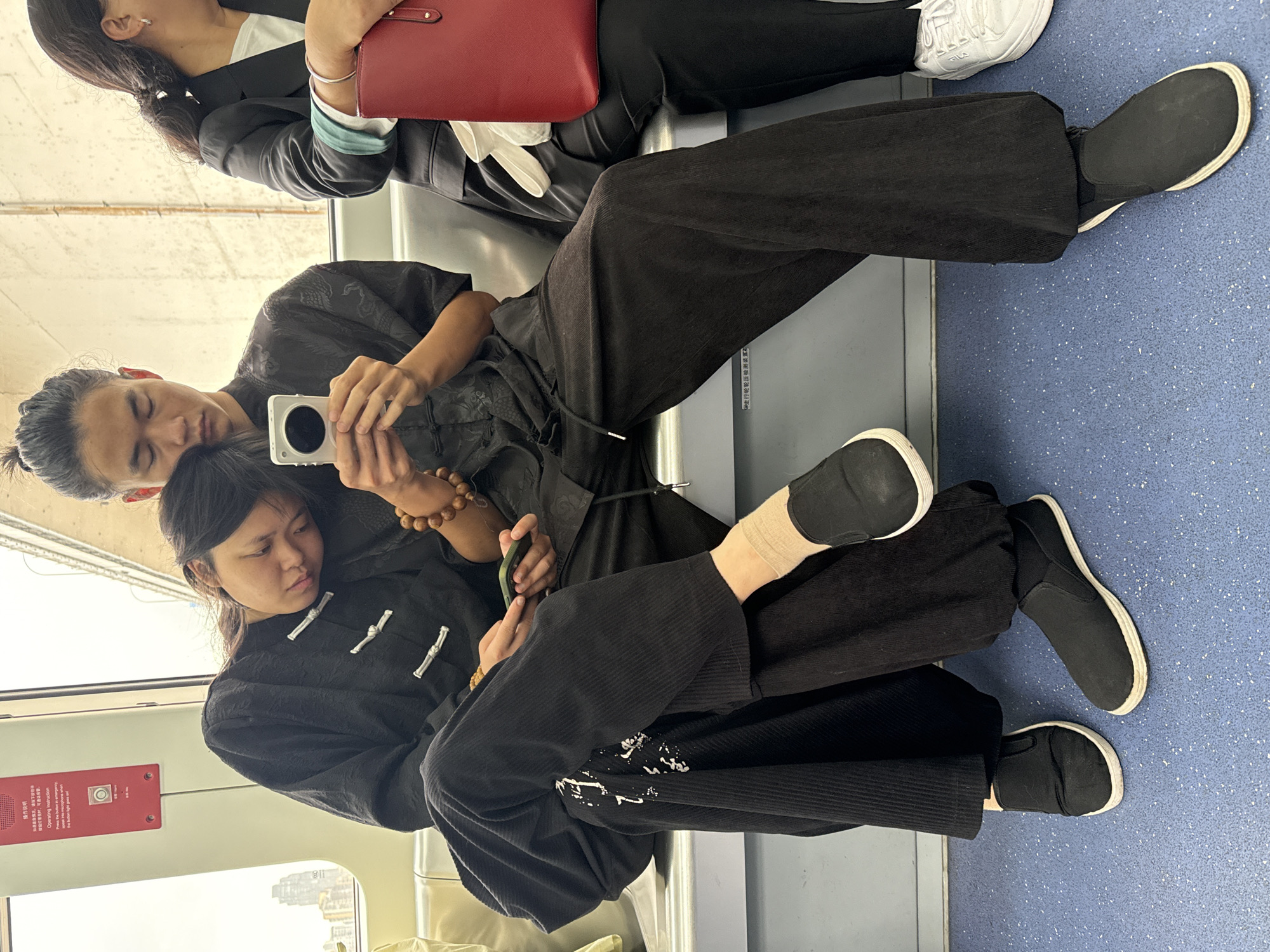The image shows a view inside what appears to be a subway train in Yuzhong District, China. Two individuals are seated, occupying multiple seats. The person closest to the camera, potentially a female, is wearing a traditional black outfit. The other person, likely a male, is taking a picture of the first person. He is wearing a black outfit. Another person is partially visible at the top of the frame wearing a white shirt, black blazer, and white shoes, holding a red purse. A window is visible with buildings visible outside. There is a red panel on the wall, and the floor of the train is blue with white speckles.
