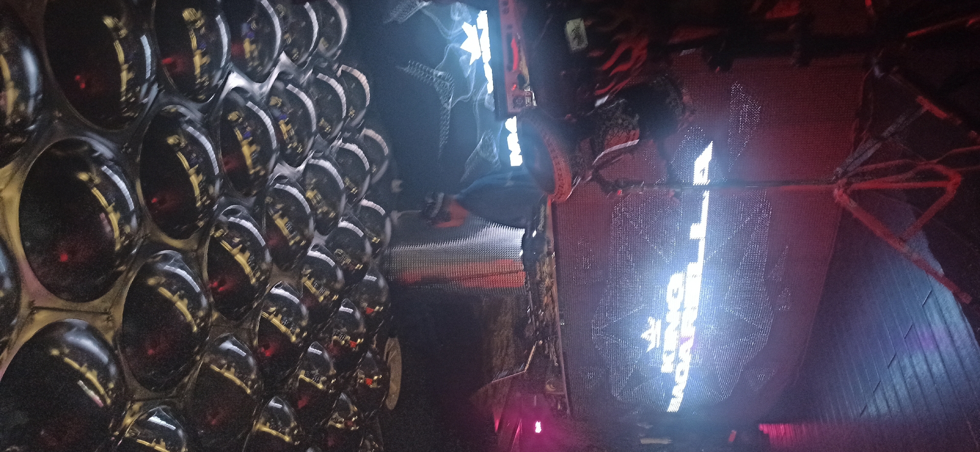 The image depicts a DJ performing at a dimly lit venue, likely a nightclub or concert hall, in Dushanbe, Tajikistan. The DJ is positioned behind a console with a large LED screen displaying bright white text that appears to read "KING BADAKHSHAN DANCE" above a logo. The ceiling is covered with numerous circular, reflective surfaces, creating a mirrored or disco-ball-like effect. Red and blue lights illuminate the scene, casting shadows and reflections on the wooden floor. The DJ, wearing a dark shirt and a baseball cap, is the central figure, with a laptop and other DJ equipment visible. The overall atmosphere suggests a lively and energetic event.