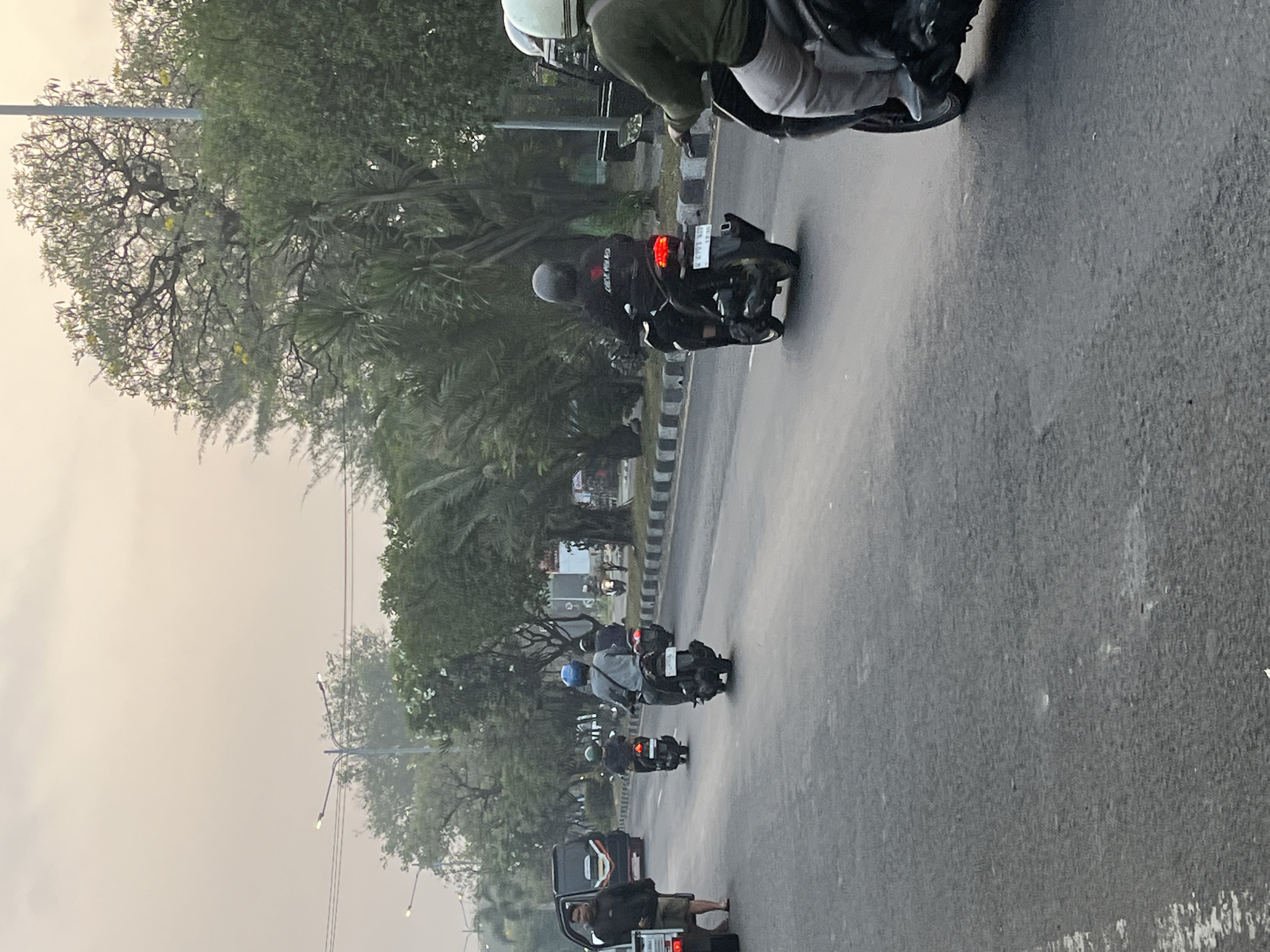 The image depicts a street scene in Kludan, Indonesia, during what appears to be early morning or late afternoon, given the soft, diffused light. The road is paved and shows signs of wear, with some patches and debris. Several motorcycles with riders are traveling in the same direction on the road. On the left side of the road, a man in a dark jacket and shorts stands near a parked black van. Further down the road, palm trees and other foliage line the street, creating a tropical atmosphere. Streetlights are visible, indicating an urban or suburban setting. The overall impression is one of ordinary daily life on a street in a warm climate.