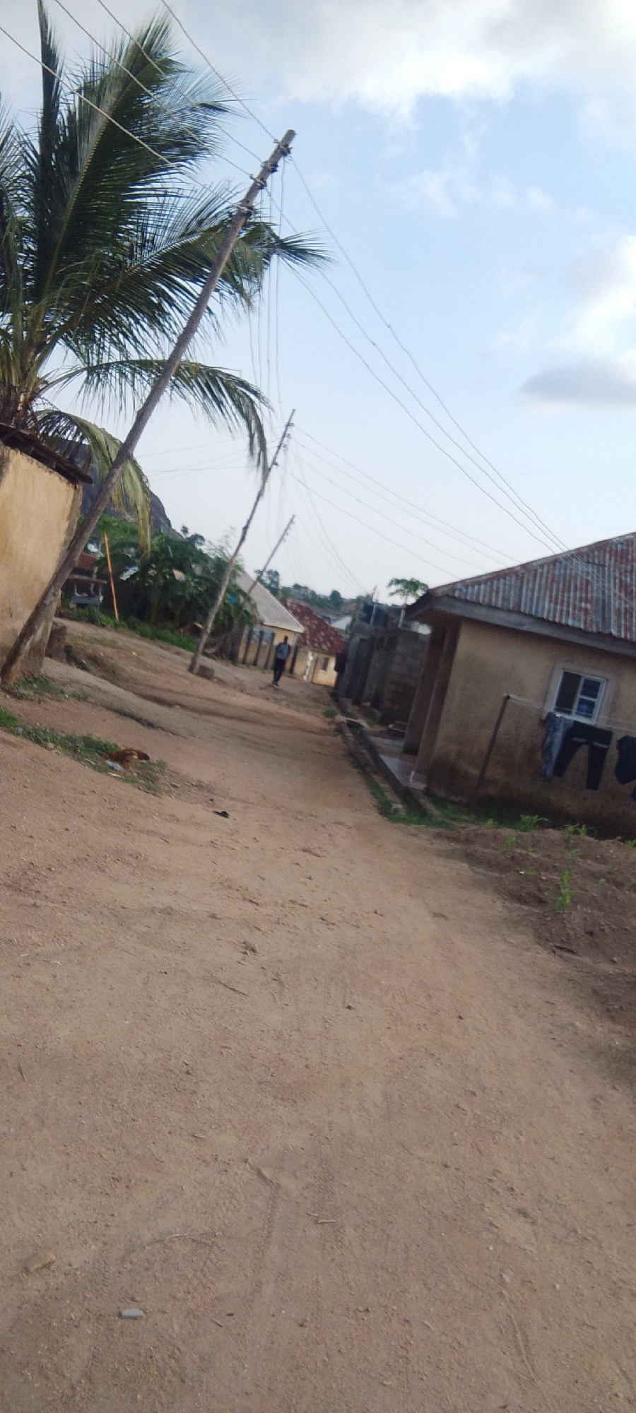 The image depicts a dirt road in an unspecified location. The road runs through a neighborhood with simple, possibly rural, houses. On the left, there's a house with a palm tree nearby. Utility poles and wires are visible overhead. In the distance, a figure appears to be walking along the road. On the right, a house has clothes hanging outside a window. The sky is partly cloudy.